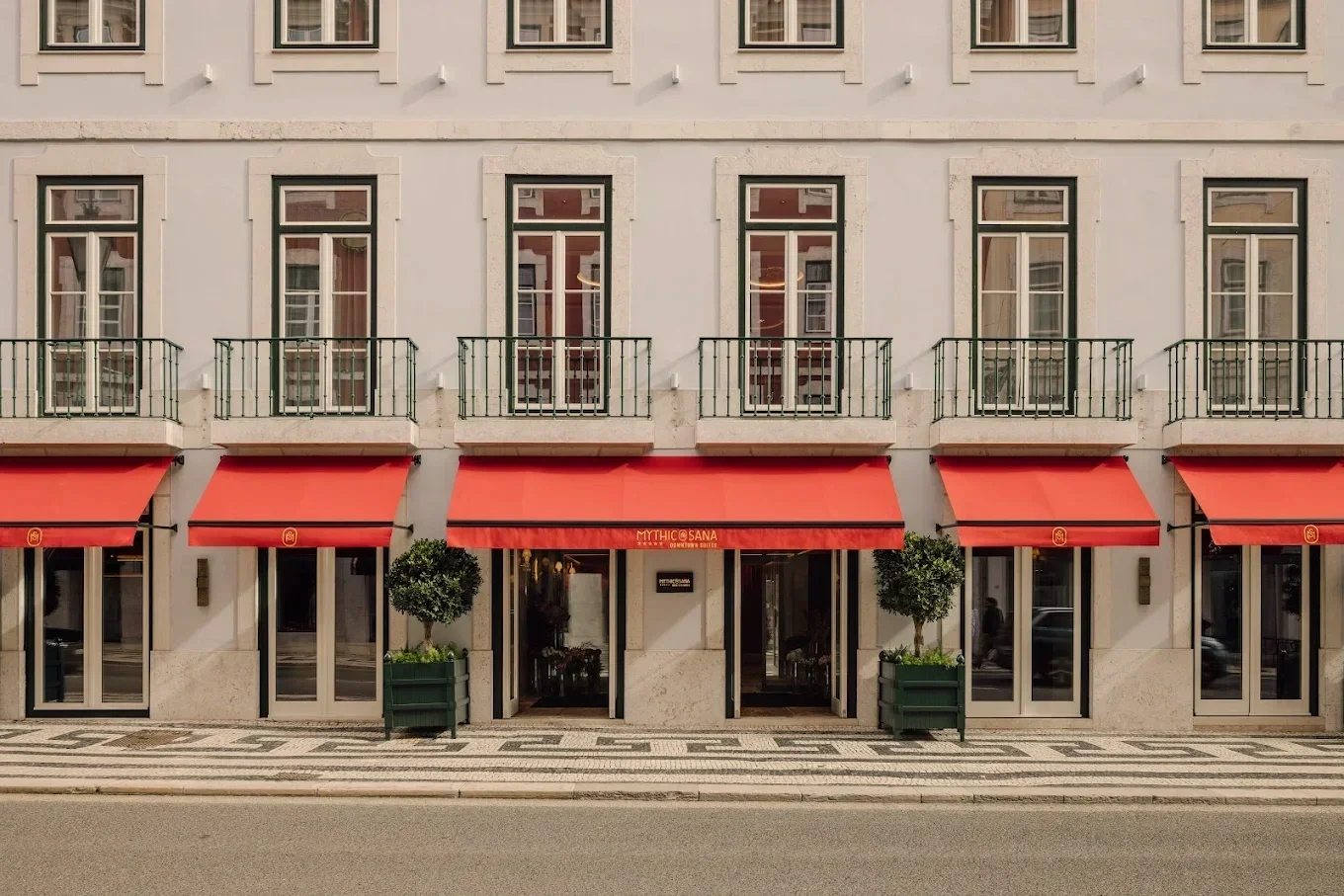 Front facade of a building with three entrances, each with a red awning and potted plants, above a patterned sidewalk.