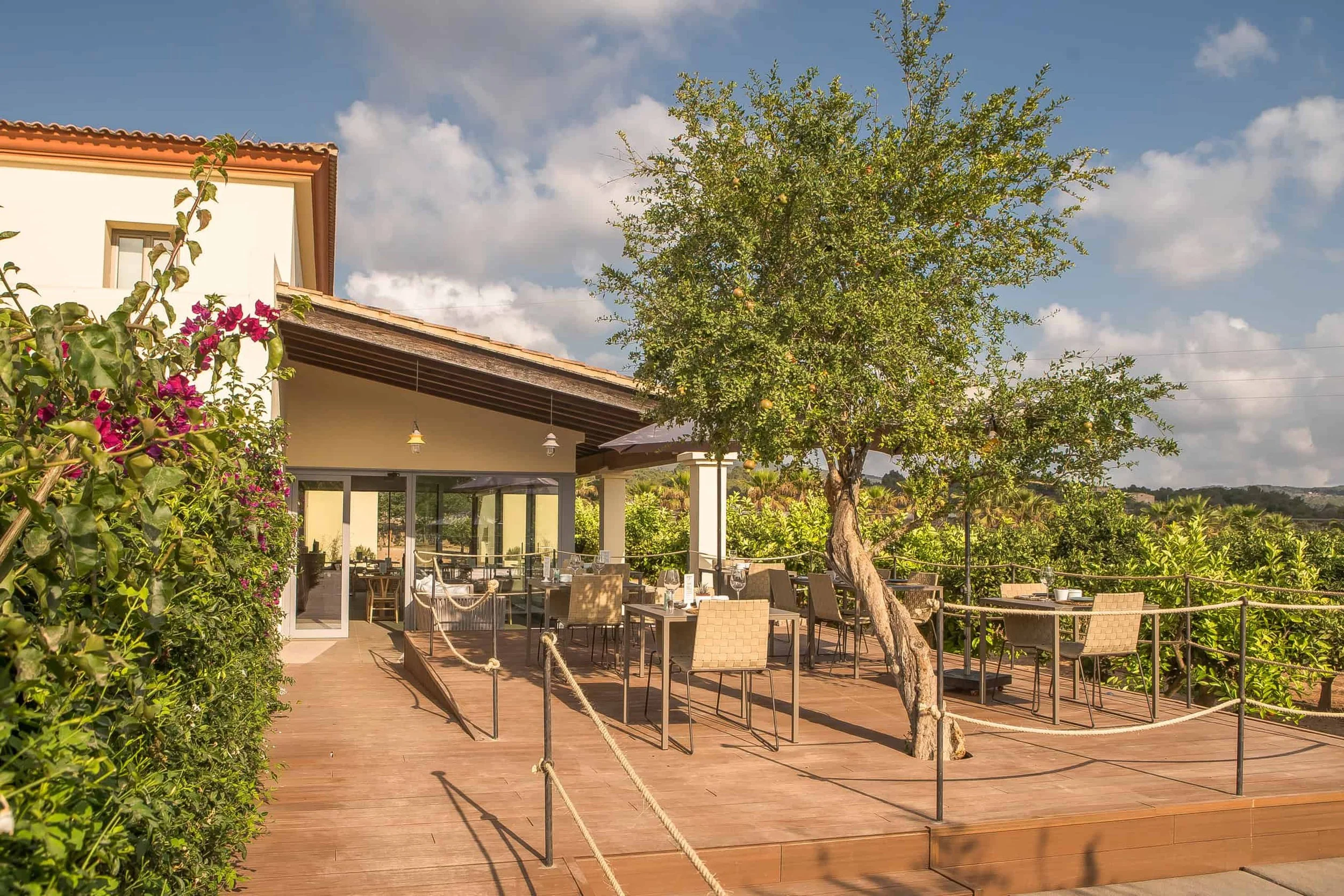 Outdoor patio area with tables and chairs, surrounded by lush greenery including a large tree and flowering bushes, with a house or restaurant building in the background under a partly cloudy sky.