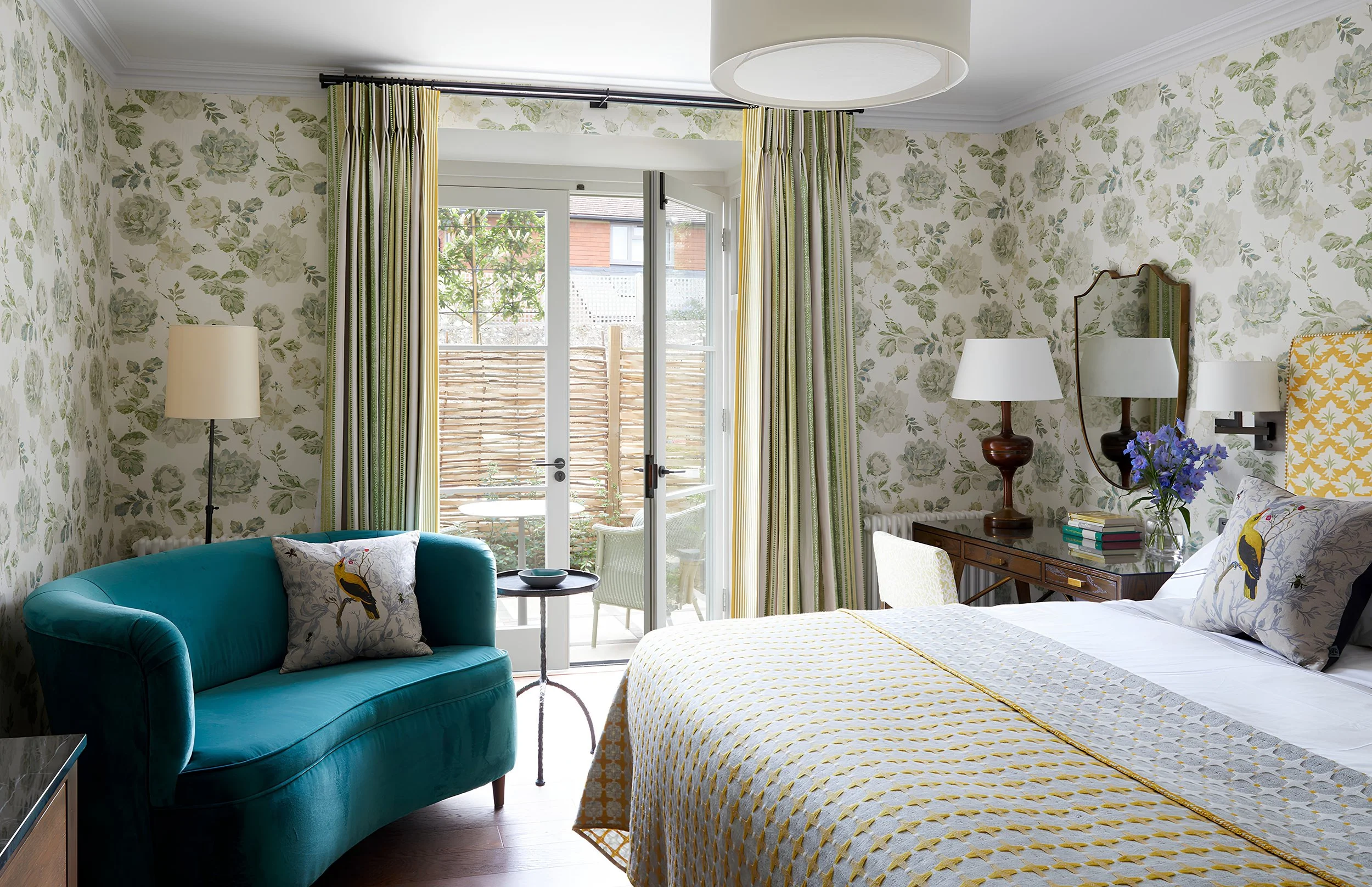 A cozy bedroom with floral wallpaper, a teal velvet sofa with a pillow featuring a bird, a small side table with a bowl, a bed with a yellow and white quilt, a wooden dresser with books and a flower vase, and a doorway leading to a small balcony with