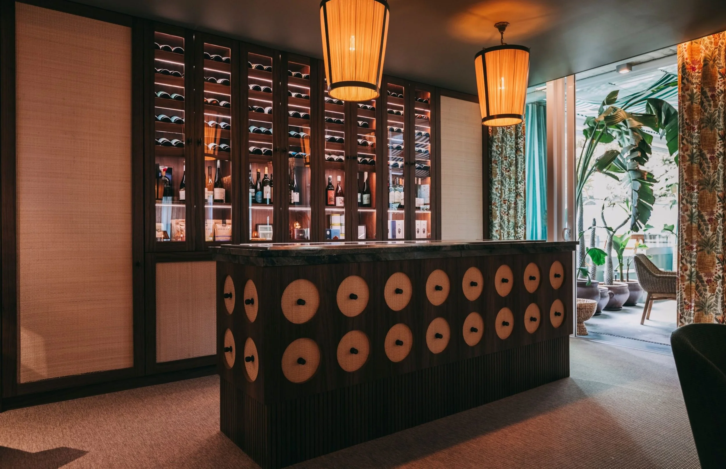 A wine bar with a dark wooden counter and a large wine rack behind it, displaying numerous bottles of wine. Two orange pendant lights hang from the ceiling, and there are large potted plants near a window with floral curtains.
