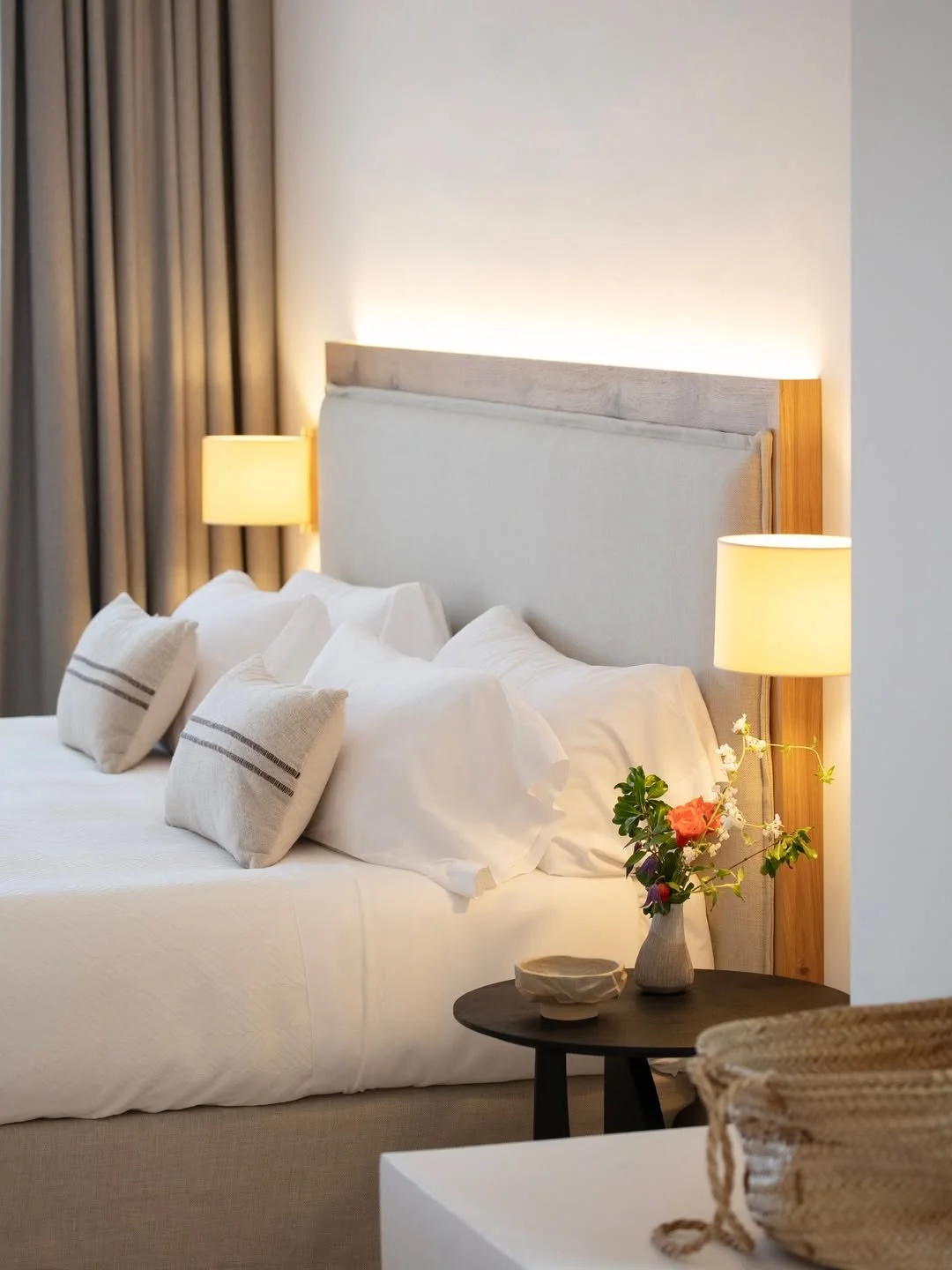 A neatly made hotel bed with white linens, multiple pillows, and a wooden headboard. Two bedside lamps are lit, and a small round table beside the bed holds a vase with flowers and a small bowl. Curtains are drawn in the background.