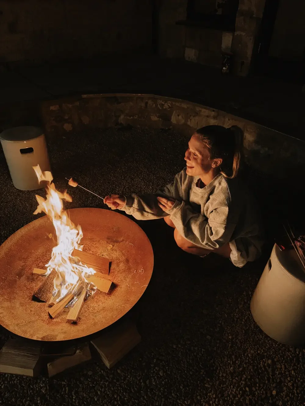 A woman is sitting near a campfire at night, roasting marshmallows on a stick, smiling.