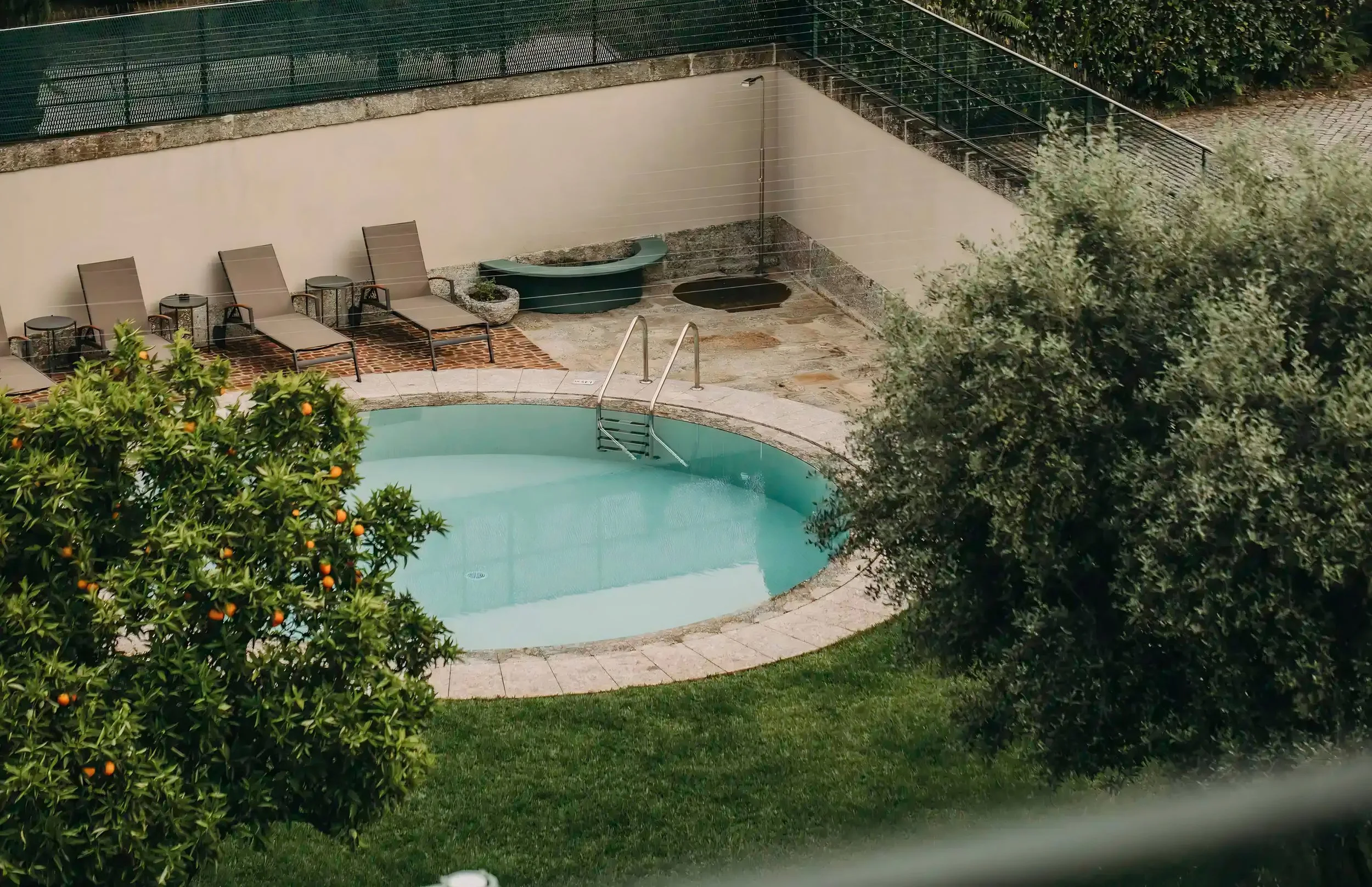 Empty swimming pool with ladder, surrounded by sun chairs, plants, and a high fence