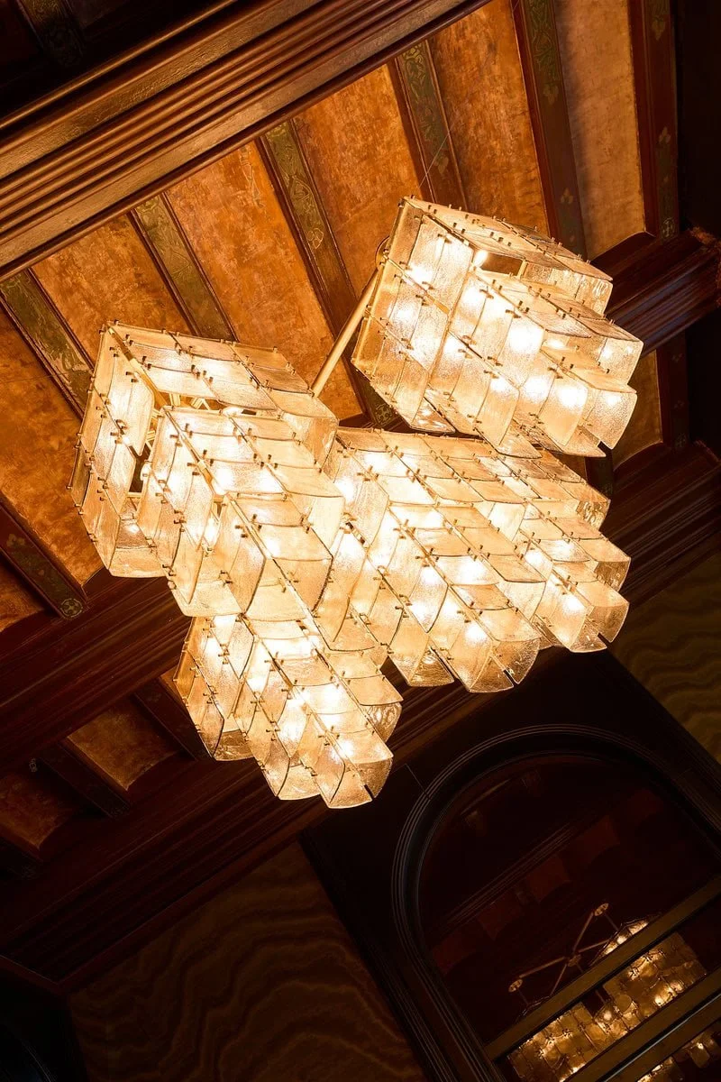 A large, ornate chandelier with multiple tiers of rectangular, frosted glass panels hanging from a decorative ceiling with wood trim and painted details.