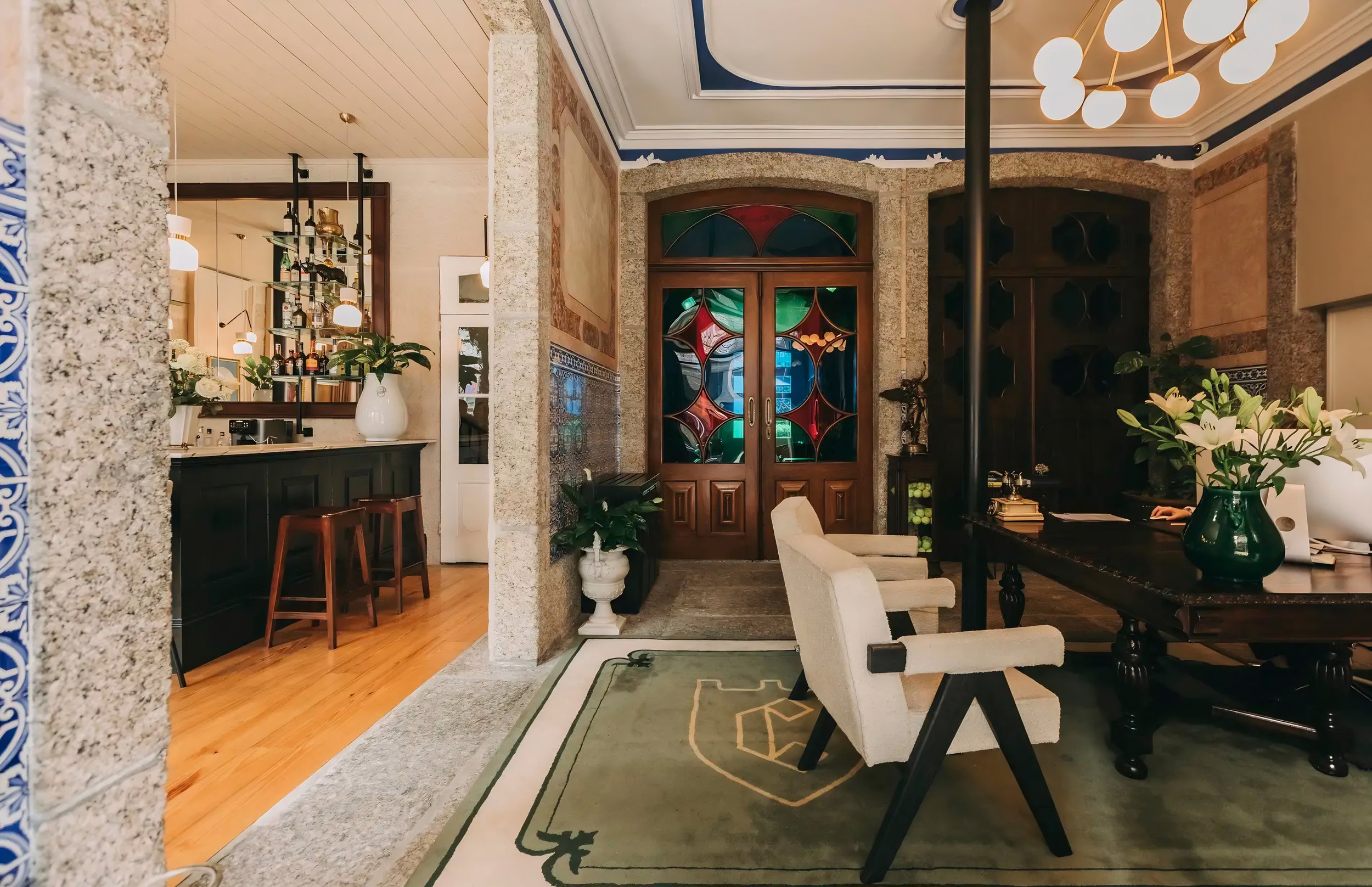 Interior of a stylish living room or lounge area with patterned rug, wooden furniture, a large dark wooden table, and floral arrangements in vases. There is a colorful stained-glass door, potted plants, and a vintage-style lamp. The walls have decora