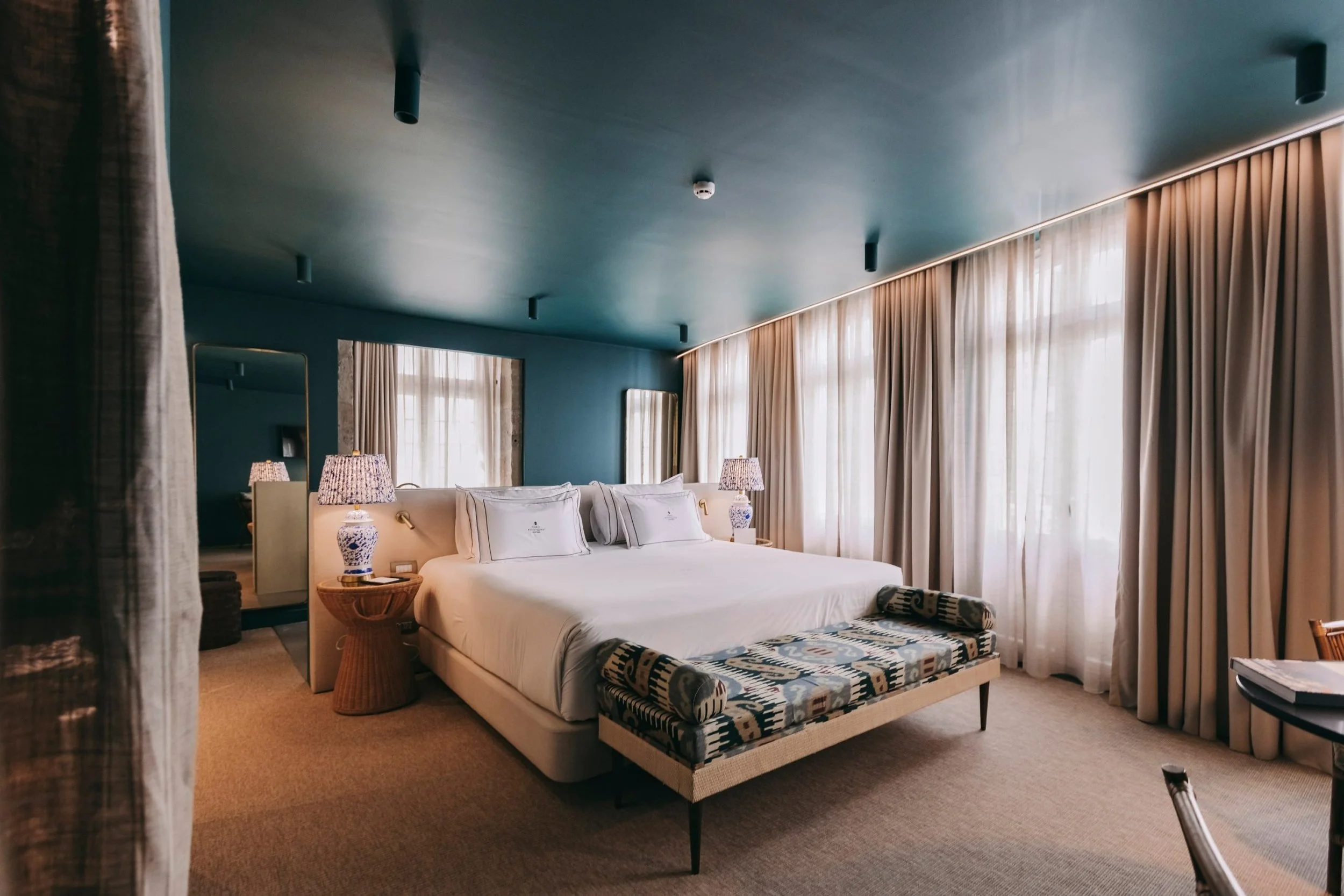 A hotel room with a large bed, bedside lamps, curtains, and large windows letting in natural light.