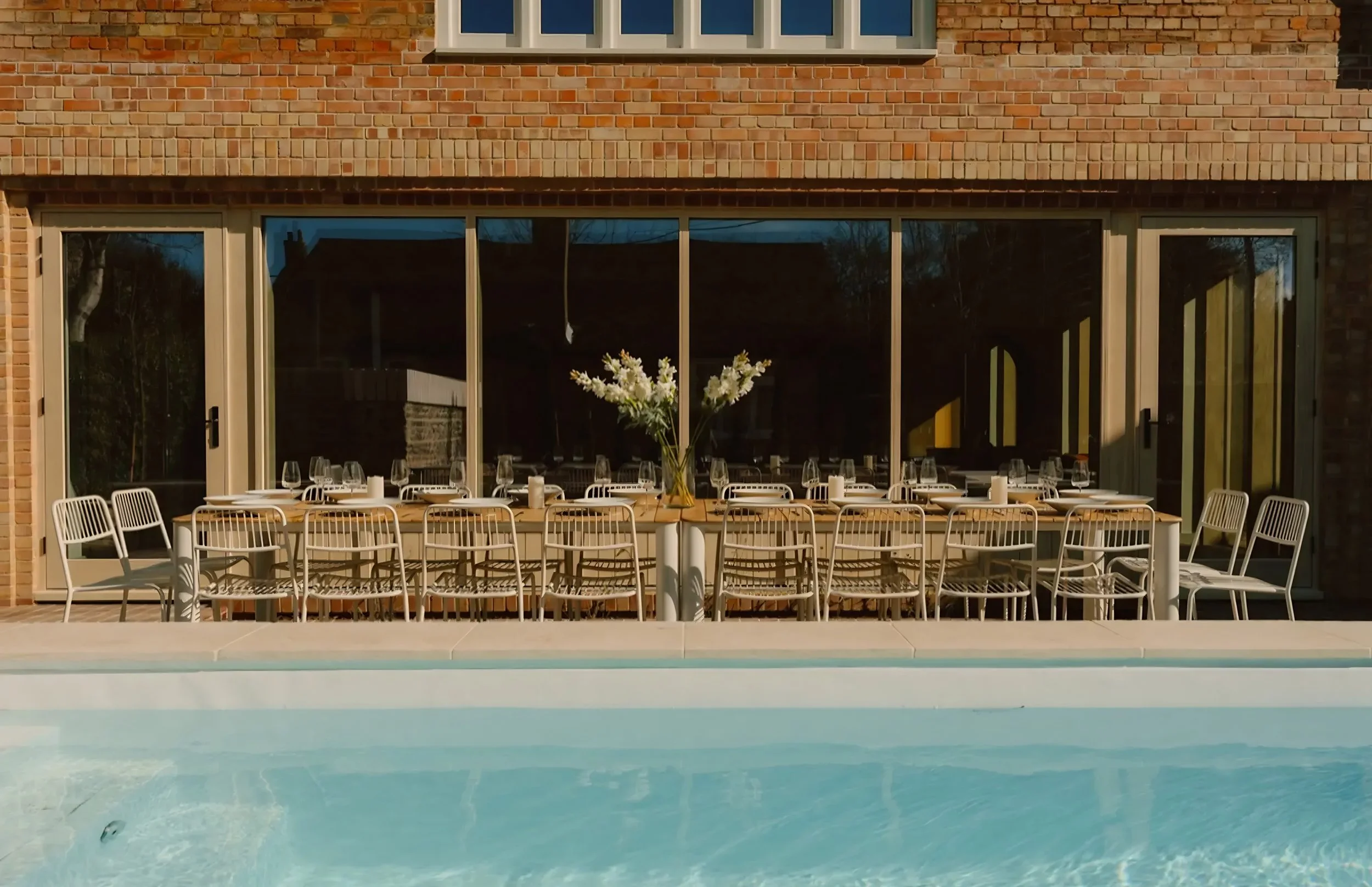 A swimming pool with blue water has a white table set with wine glasses and candles, surrounded by white chairs, in front of large glass doors leading into a brick house.