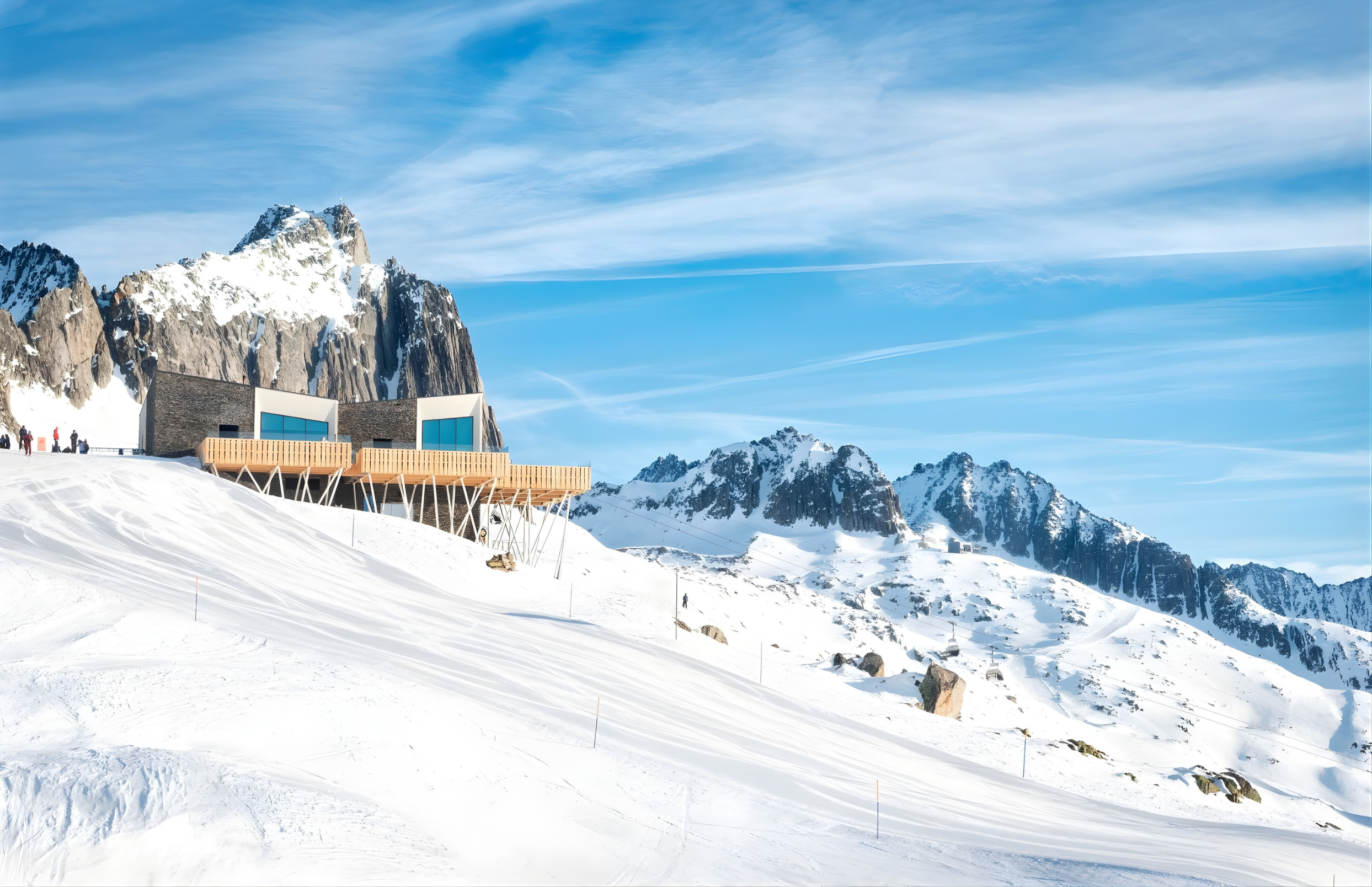 A modern building with large windows on a snow-covered mountain slope, with a rugged snowy mountain range and blue sky in the background.