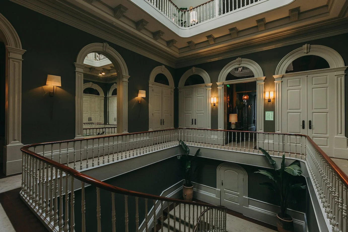Interior view of a luxurious, multi-story building with dark green walls, white decorative moldings, and a balcony with a white and wood railing. There are several wall-mounted lamps providing warm lighting, and large arch-shaped windows and doors wi