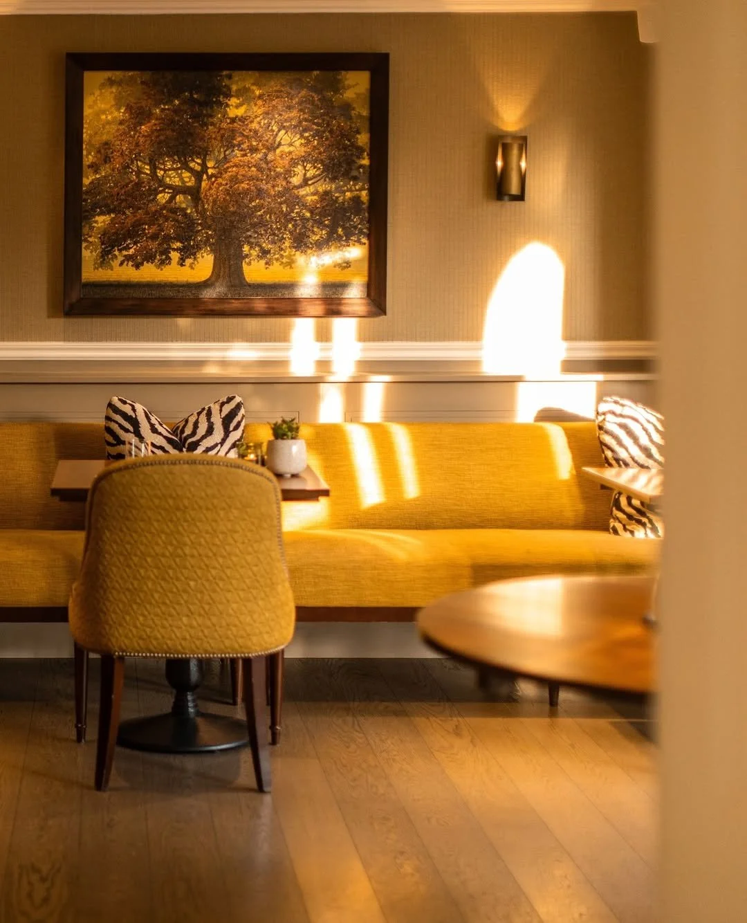 A cozy dining area with a yellow upholstered bench, a single zebra print pillow, a wooden table, a yellow chair, and a small potted plant. A large framed picture of a tree hangs on the beige wall, and sunlight streams in, creating bright patches of light and shadow.
