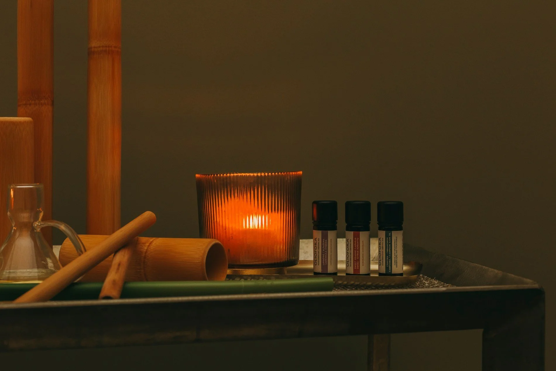 A dinner table with essential oils, a glass candle holder with a lit candle, wooden rolling pins, glass beaker, and a tray with candles and herbs, set against a dark background.