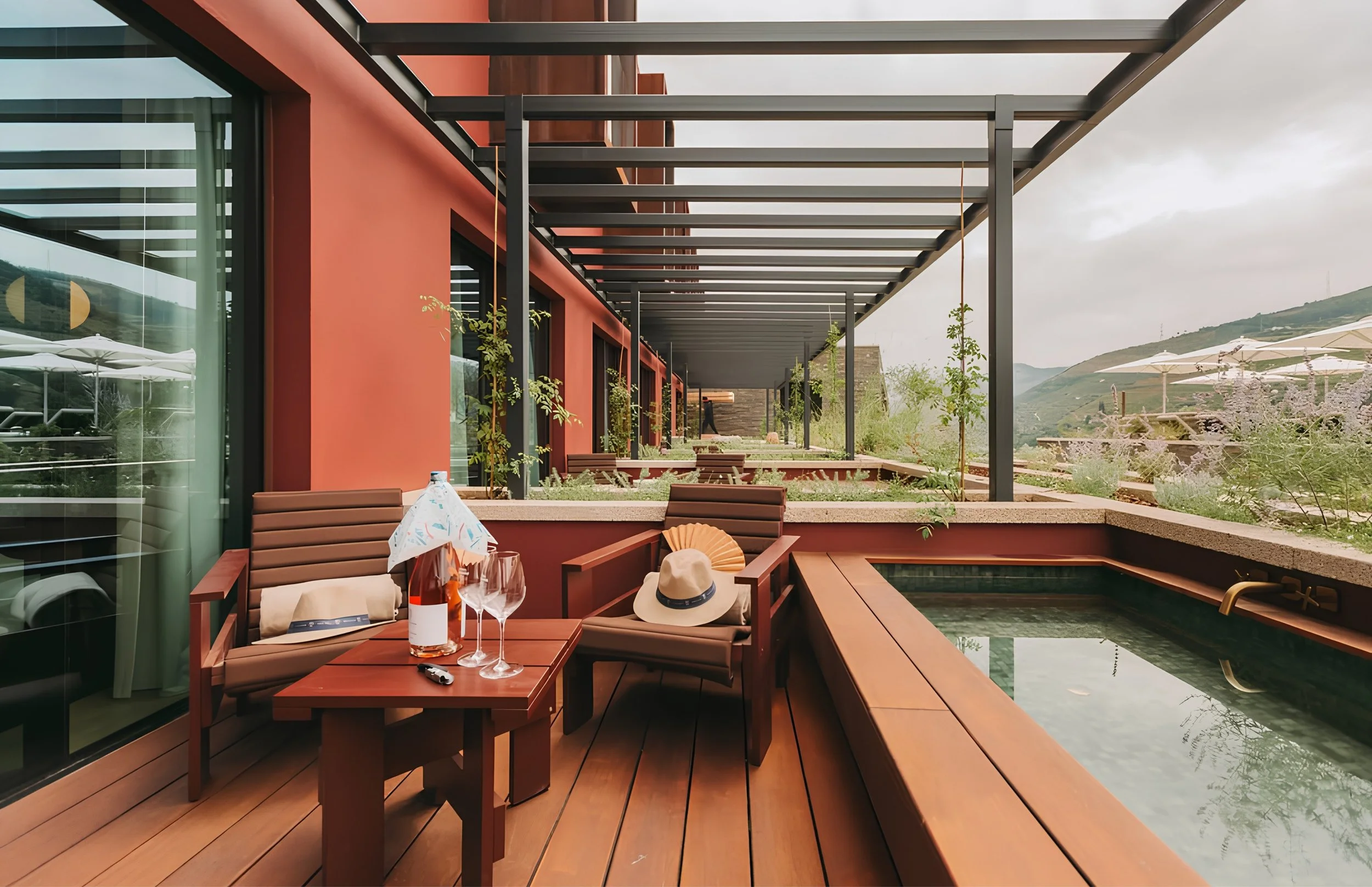 Outdoor dining area with wooden tables and chairs, some with drinks and accessories, e greenery and mountains in the background, partially covered by a modern pergola with horizontal beams.