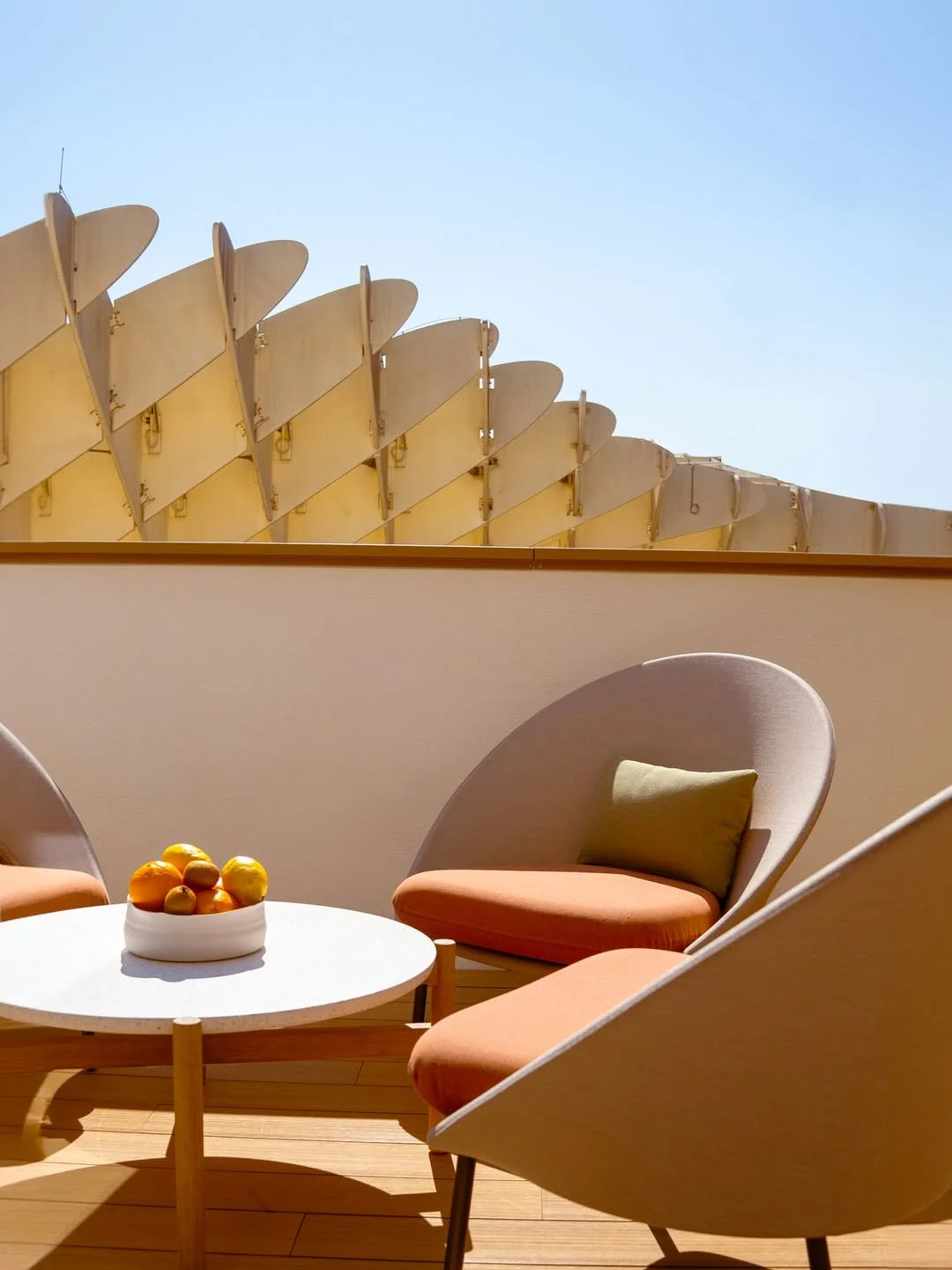 Outdoor seating area with modern chairs and a small round table holding a bowl of fruit, against a modern architectural backdrop with a blue sky.
