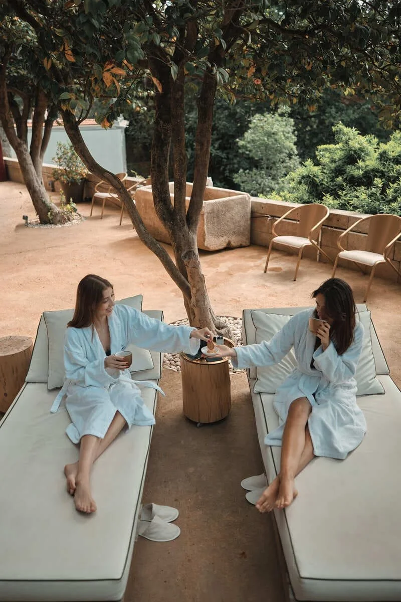 Two women in white robes relaxing outdoors on lounge chairs, sharing a drink under a large tree with greenery in the background.
