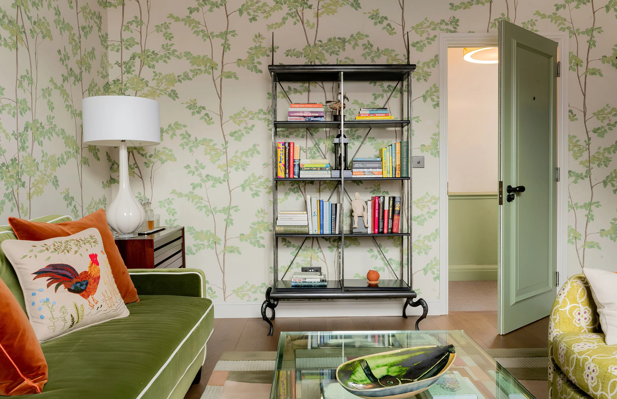 Living room with green velvet sofa, colorful pillows, a glass coffee table, a tall bookshelf with books and decorative items, a white table lamp, and wallpaper with green leafy branches. A door is open to another room with a modern ceiling light.