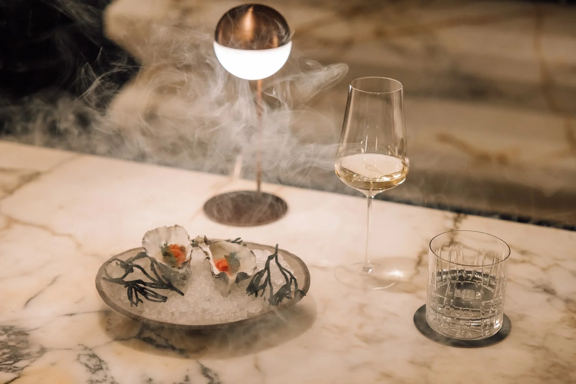 Elegant restaurant table setting with a bowl of oysters on ice, a glass of white wine, an empty wine glass, and a crystal tumbler, with a dark, textured background.