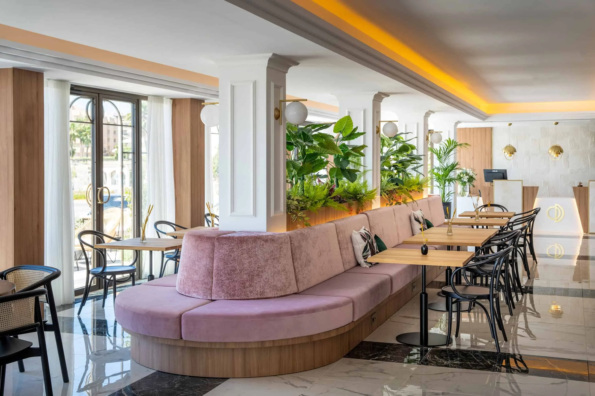 Modern indoor dining area with pink curved booth seating, wooden tables, black chairs, large plants, and large windows with white curtains.