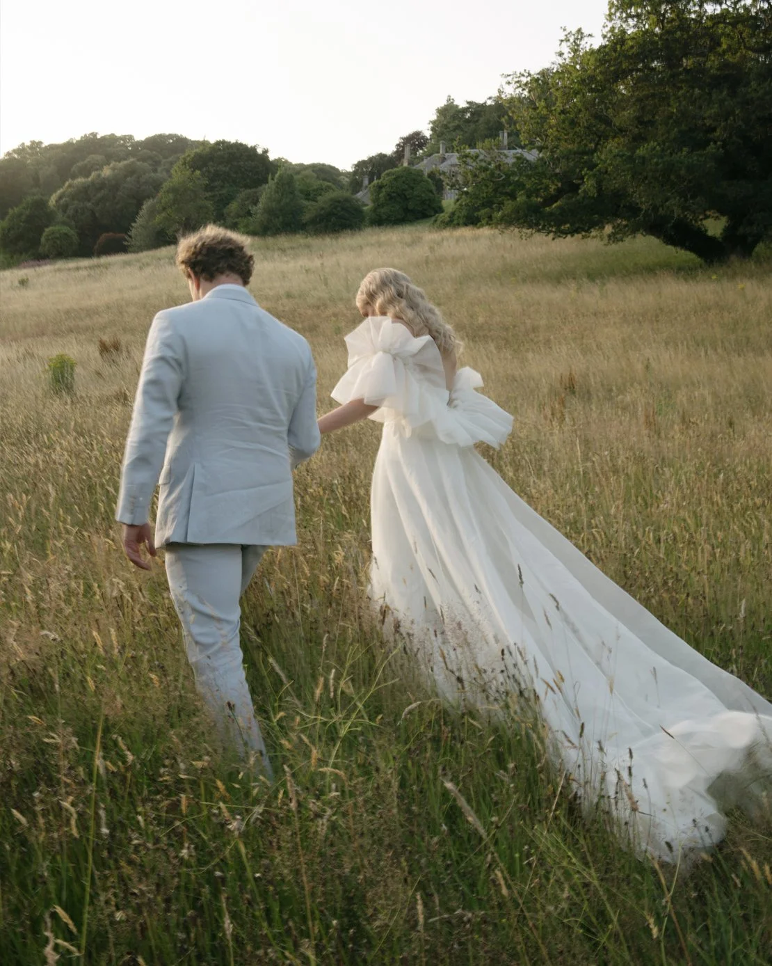 A couple dressed in wedding attire walking through a grassy field at sunset, with the bride in a long white gown with large bows on her sleeves, and the groom in a light-colored suit.