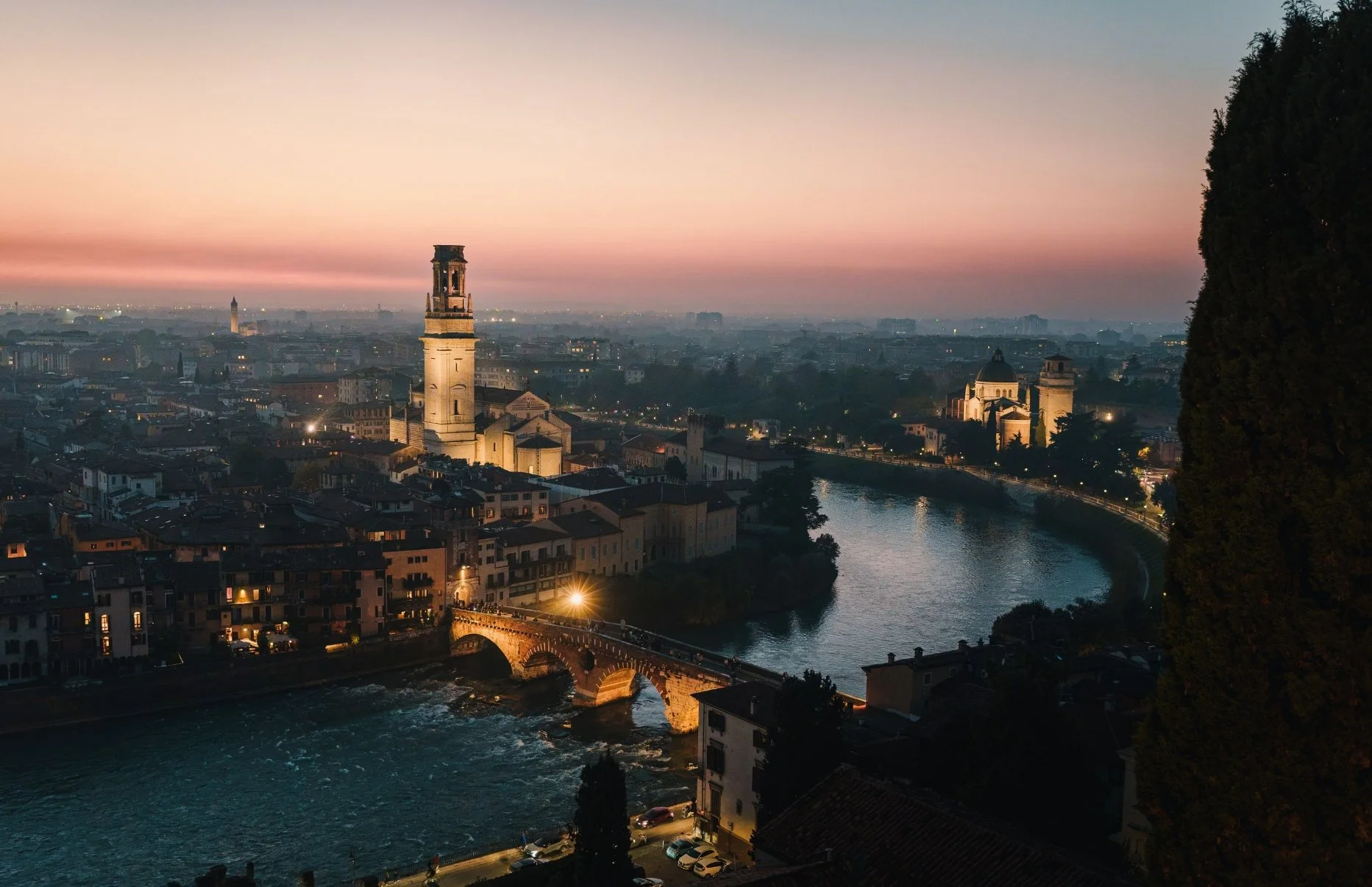 Cityscape of northern Italy at dusk, featuring the Adige River, historic bridges, and prominent bell towers and buildings illuminated along the riverbank, with a colorful sky in the background.