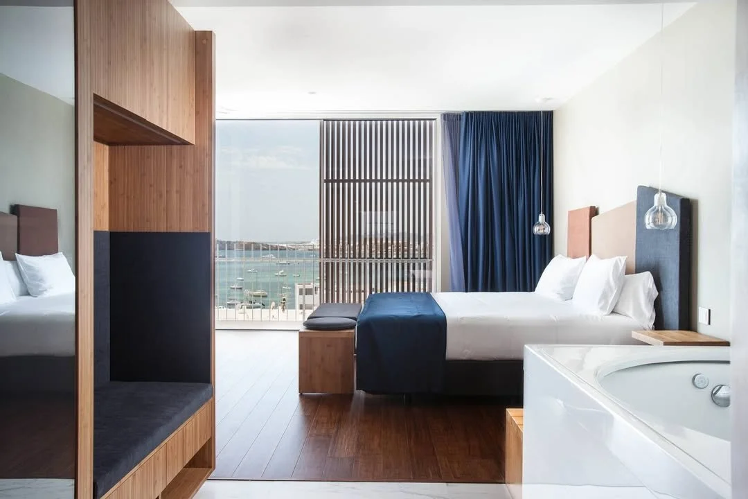 Modern hotel room with a large bed, wooden furnishings, and a view of a marina through large windows with blue curtains.