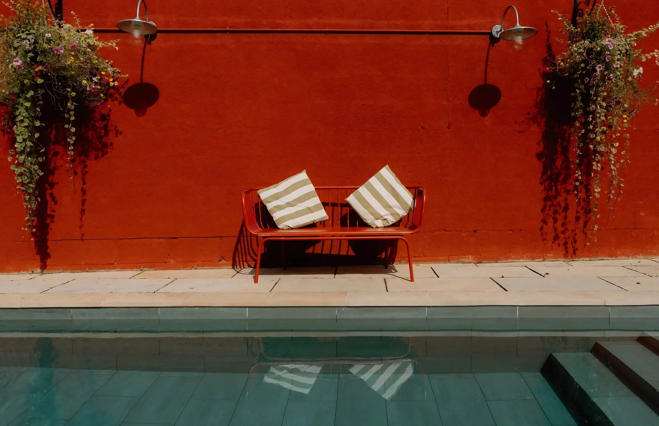 A red wall with two metal outdoor wall lamps casting shadows. Hanging on the wall are two overflowing flower baskets with pink and purple flowers and greenery. In front of the wall, there is a metal bench painted red with two striped cushions, one be