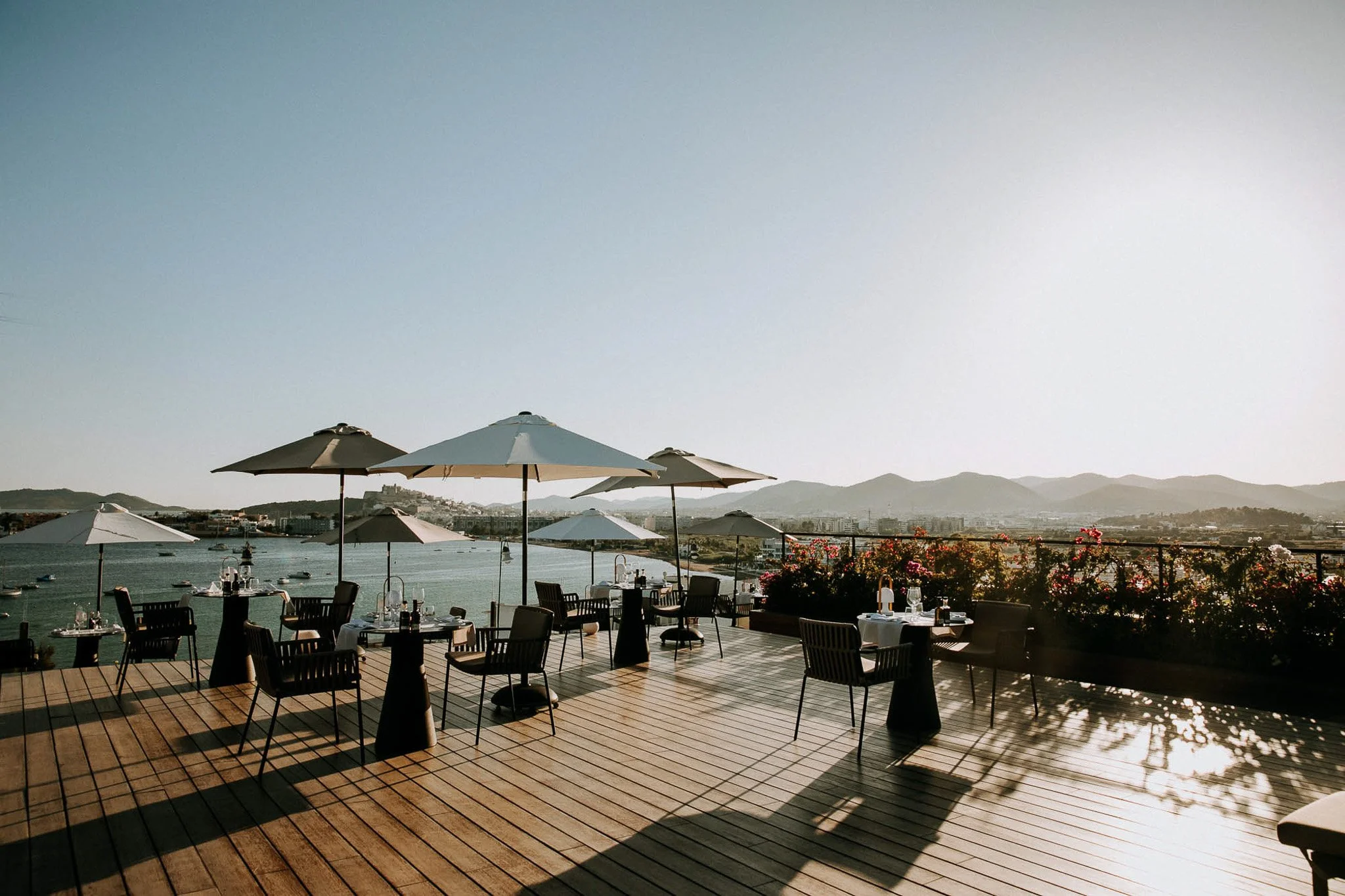 An outdoor restaurant with tables, chairs, umbrellas, and a view of a lake, mountains, and a clear sky in the background.