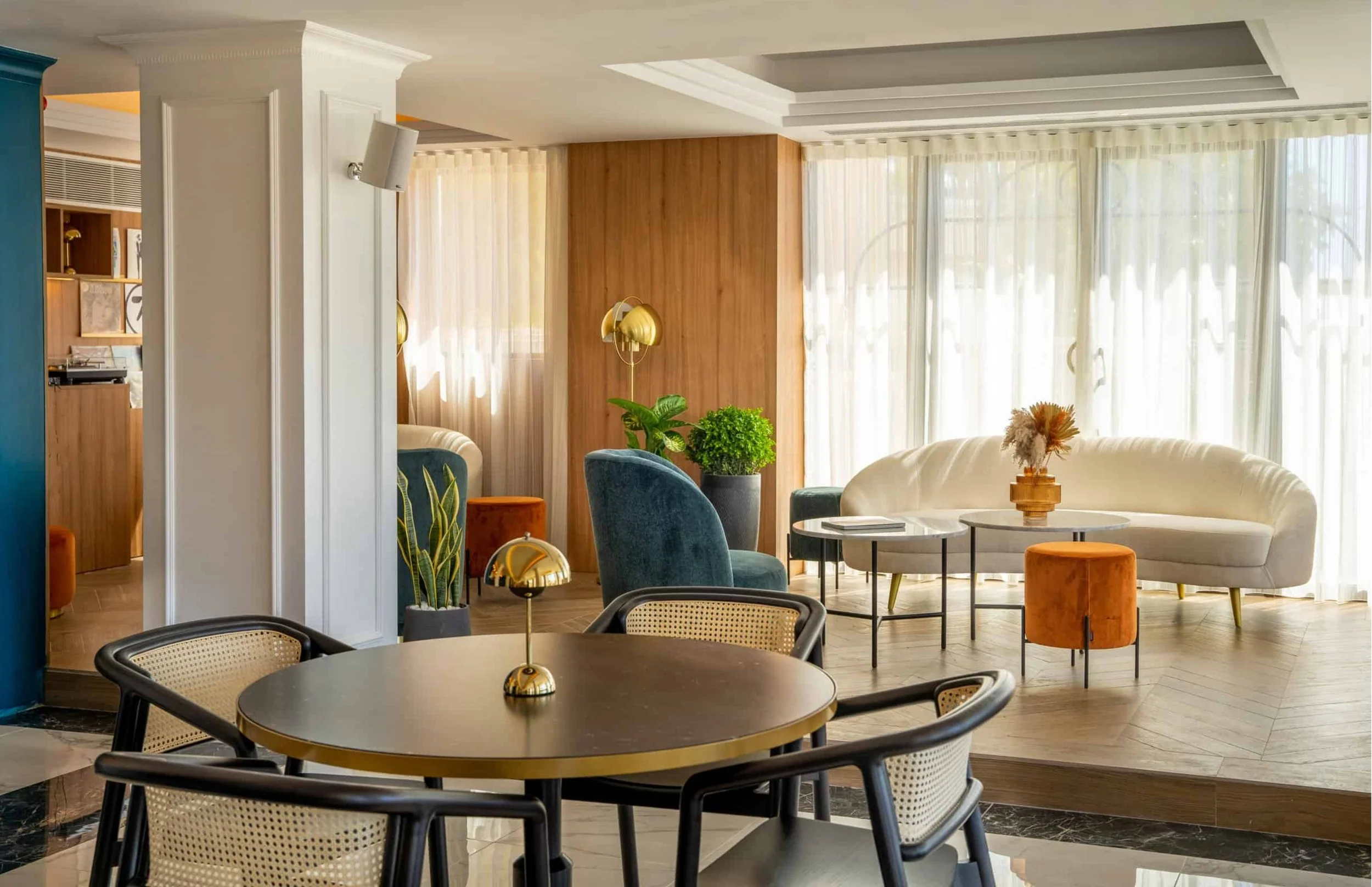 A spacious and airy living room with large windows covered by sheer curtains, a white curved sofa with an orange throw pillow, blue armchairs, round coffee tables, and decorative plants, illuminated by natural sunlight.