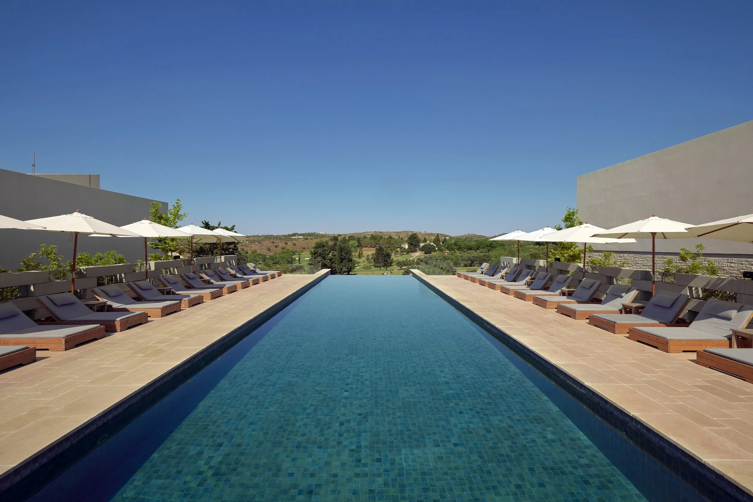 Outdoor swimming pool with lounge chairs and umbrellas, set against a backdrop of green landscape and hills under a clear blue sky.
