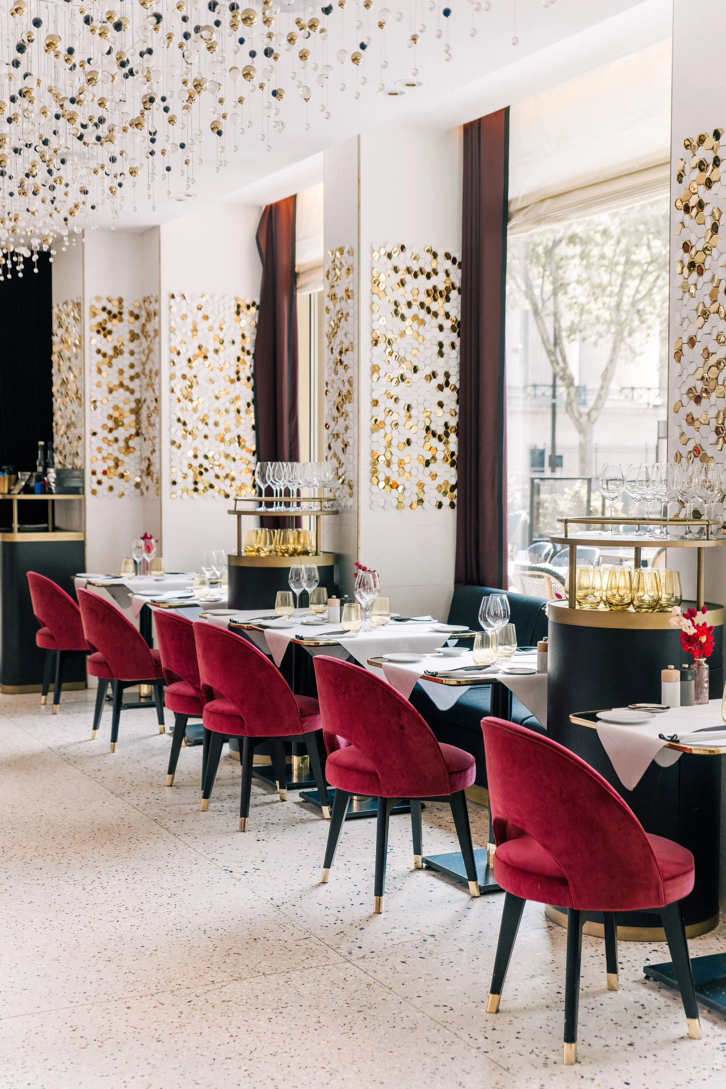 Inside a modern restaurant with pink velvet chairs, white tablecloths, and wine glasses. The walls are decorated with gold and white hexagon tiles, and large windows with beige and maroon curtains allow natural light to fill the space.