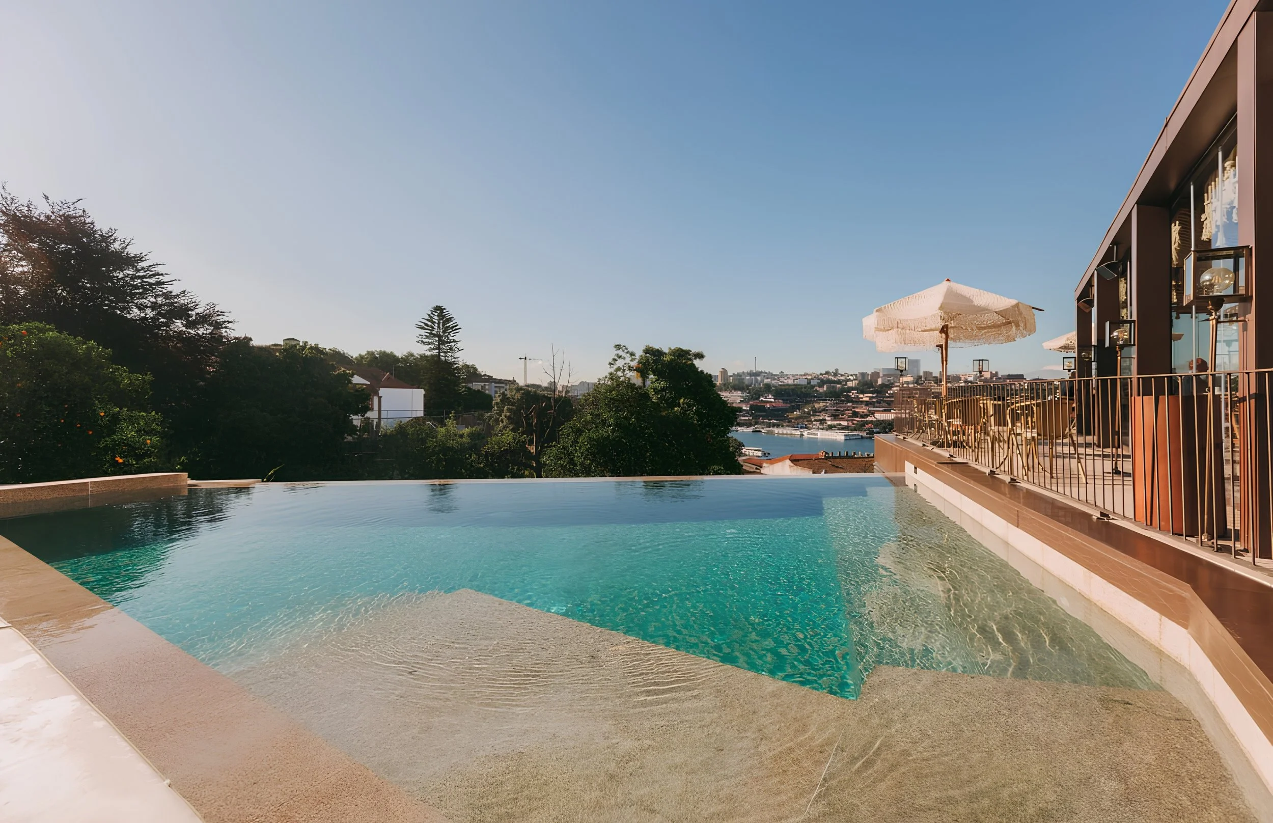 A rooftop infinity pool with a city skyline view, outdoor seating with umbrellas, and lush greenery in the background.
