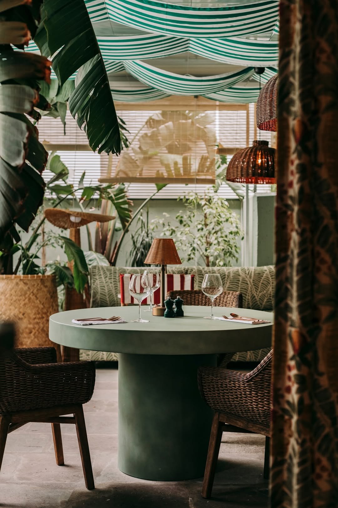 A cozy indoor restaurant corner with a round green table set with two wine glasses, cutlery, and a small lamp. Behind, there is a sofa with patterned cushions and lush indoor plants. Decorative fabric drapes hang from the ceiling, creating a warm ambiance.