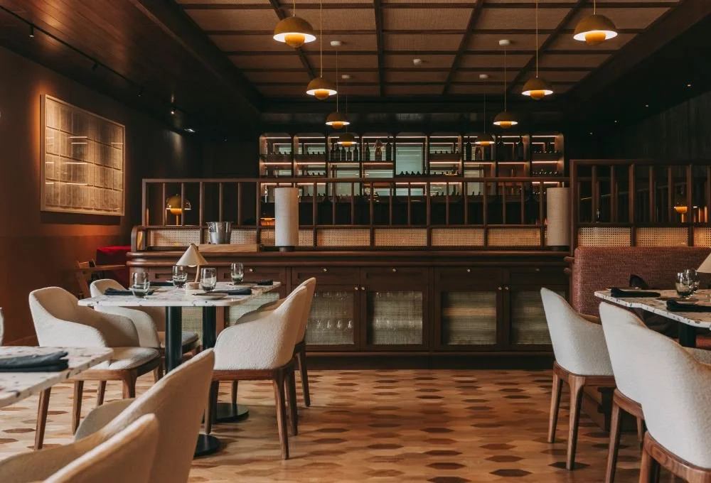 Interior of a modern restaurant with wooden decor, round tables set with napkins, glasses, and cutlery, beige upholstered chairs, warm ambient lighting, and a bar area in the background.
