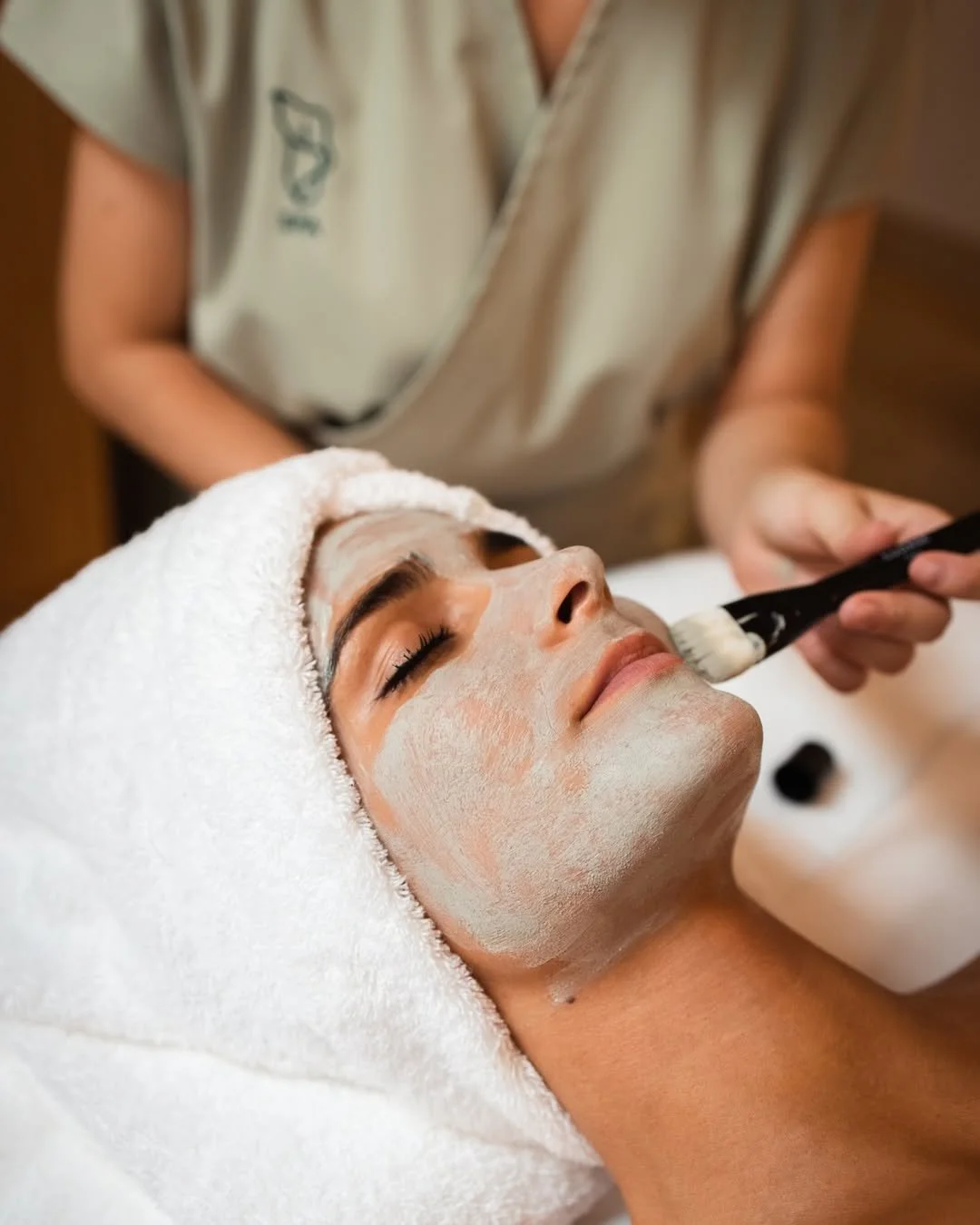 A person receiving a facial treatment, lying down with a towel wrapped around their head, while a skincare professional applies a mask using a brush.