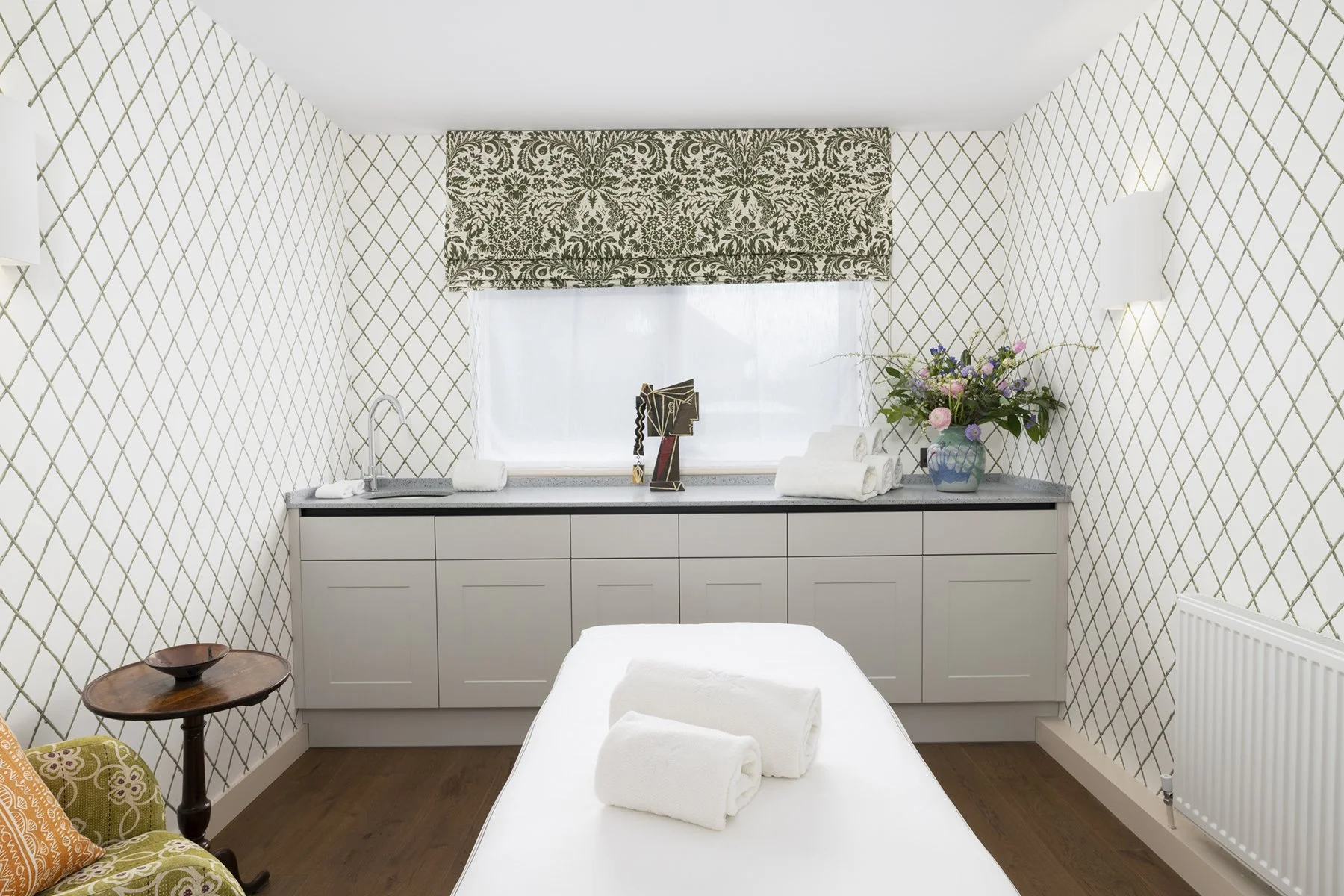 Spa or massage room with a bed in the foreground, white towels on top, a side table with a bowl, patterned wallpapered walls, a large window with a dark patterned valance, a cabinet counter with a sculpture and towels, a flower vase with flowers, and wall-mounted lamps.