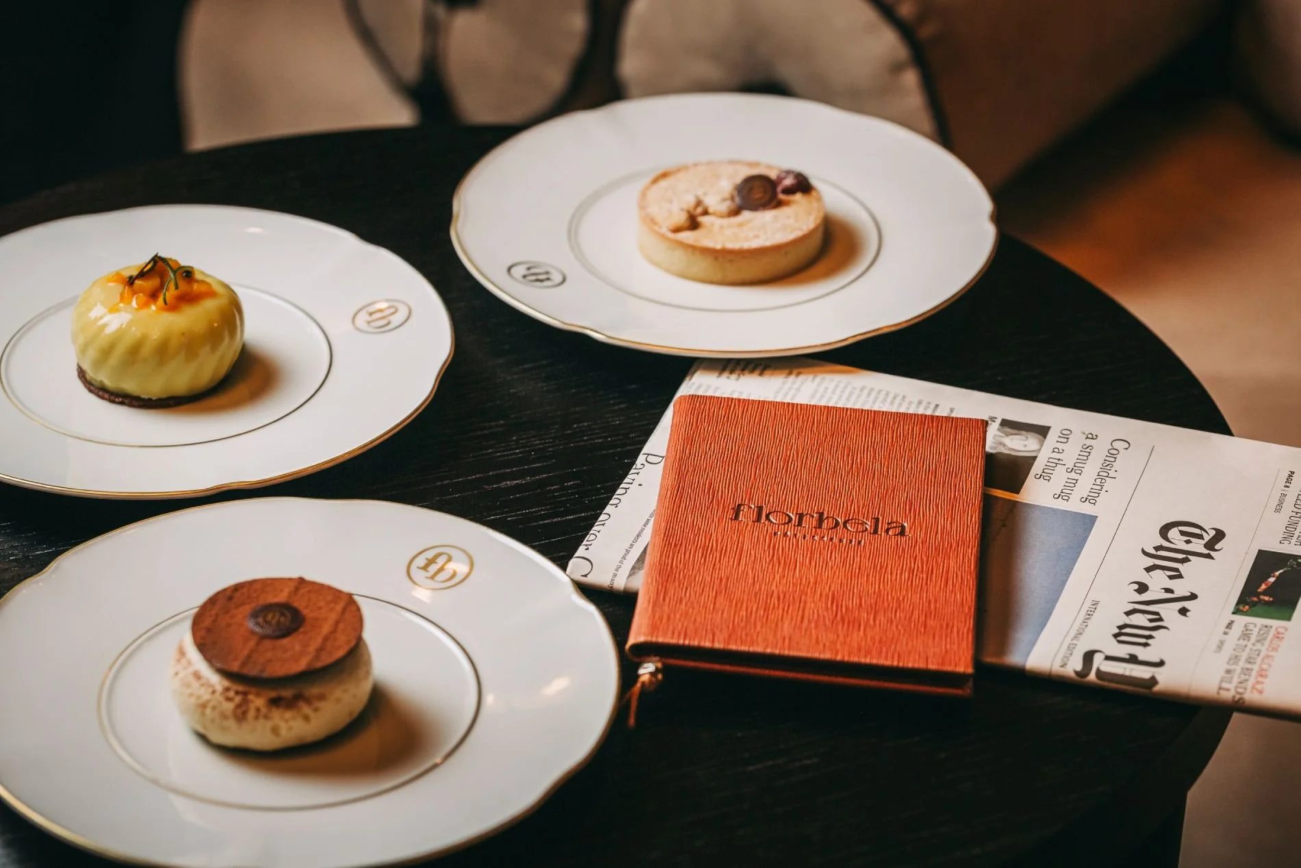 Various desserts on elegant white plates with gold trim, a closed orange Hermès notebook, and a newspaper on a dark wooden table.