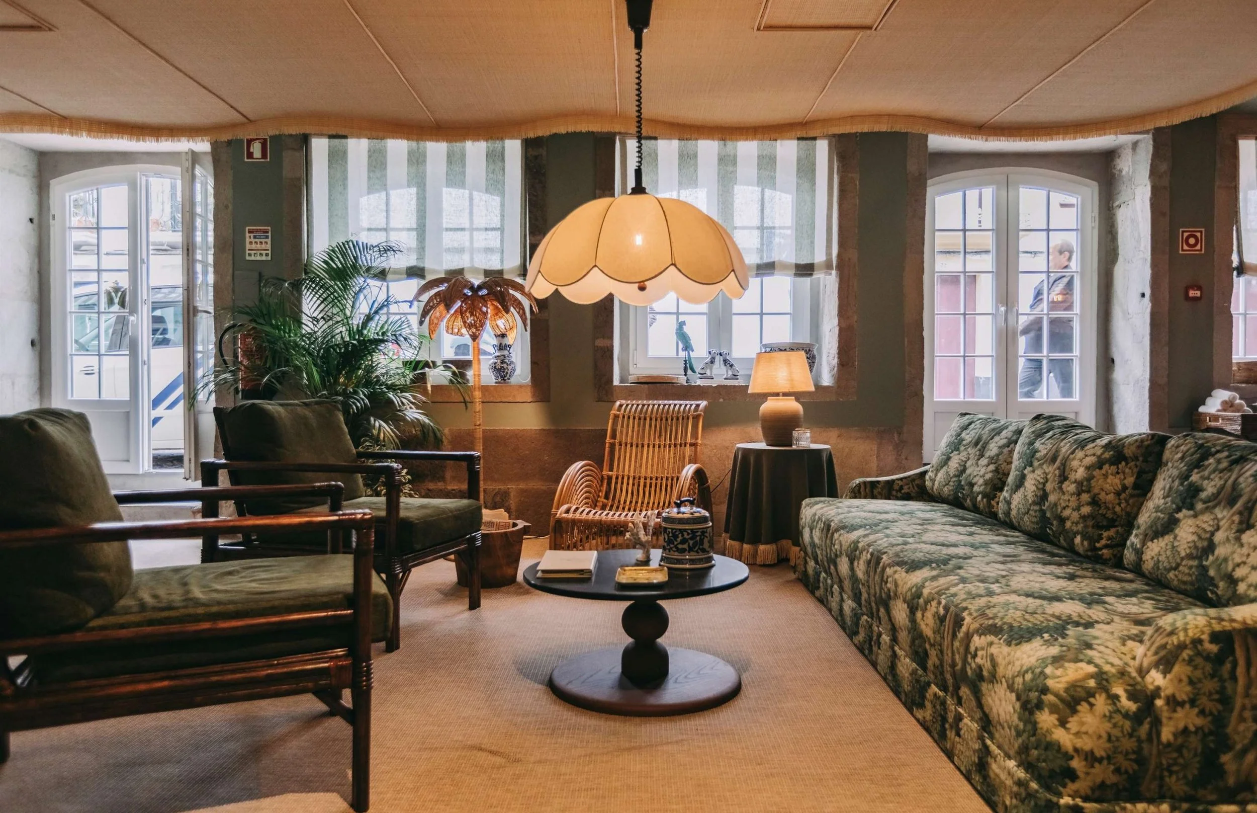 A cozy living room with large windows, patterned sofa, wooden armchairs, multiple lamps, a round coffee table with books and decorative items, and indoor plants.