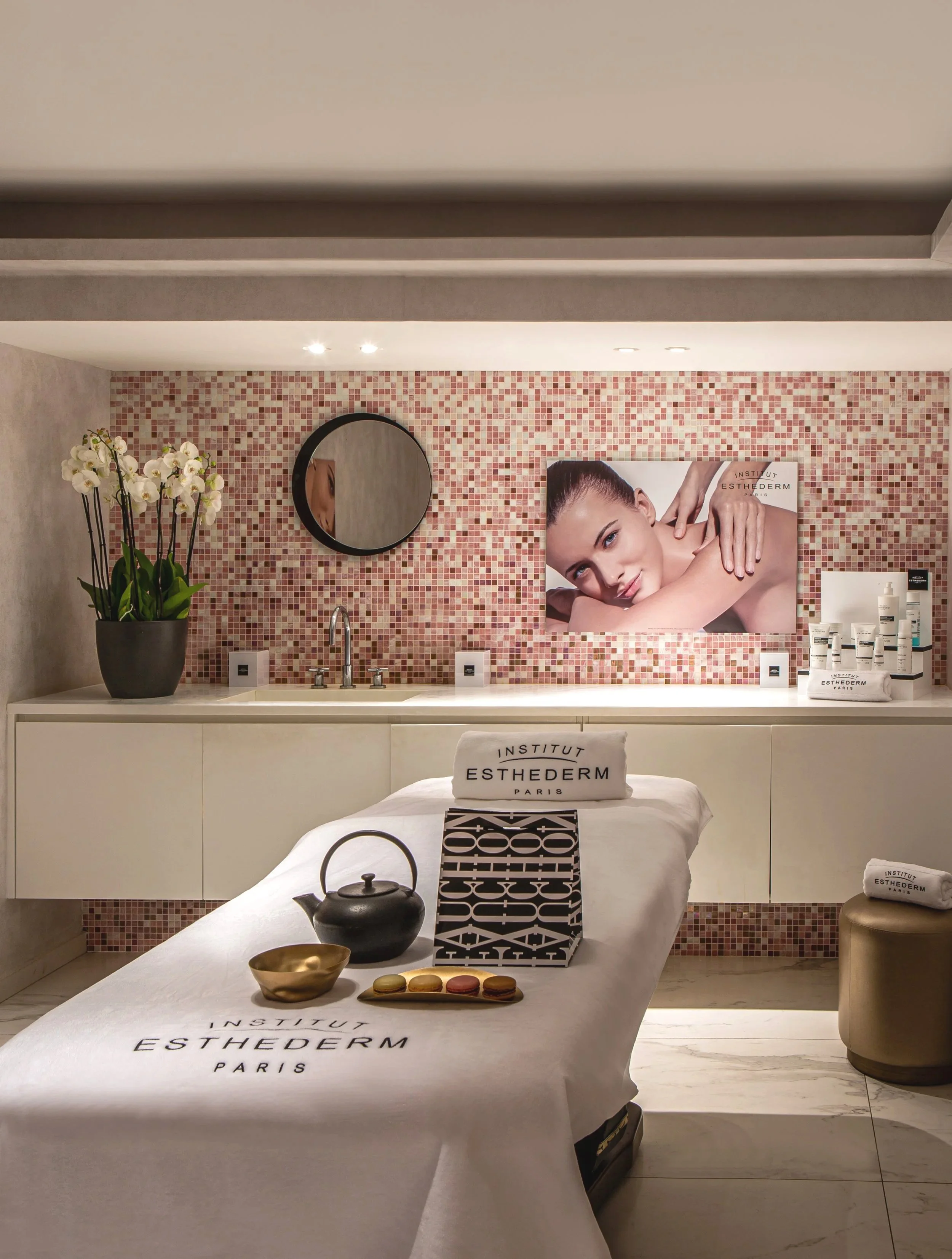 A wellness or spa room with a treatment table covered by a white cloth with the words 'Institut Esthederm Paris.' The table has a black teapot and bowls with colorful macarons. In the background, there is a mosaic pink-tile wall, a large poster of a woman receiving a massage, a round mirror, a potted orchid plant, and a counter with skincare products.
