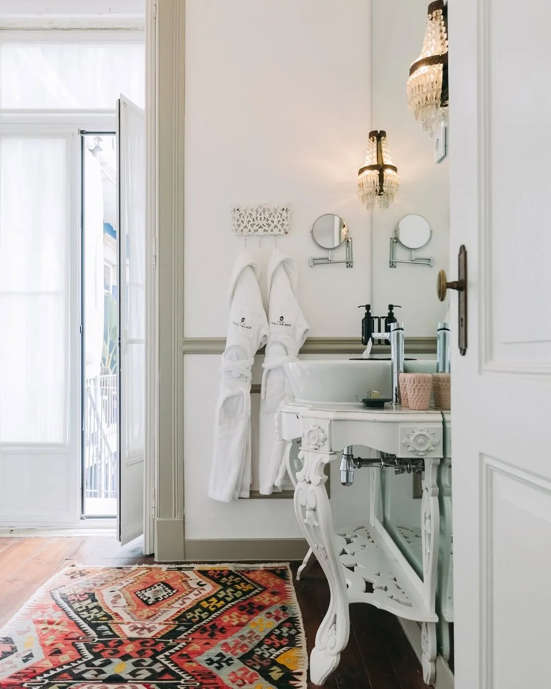 A cozy white bathroom with a vintage vanity, two small wall mirrors, and a decorative chandelier. Towels hang on a wall hook beside the door, and a colorful rug covers part of the wooden floor.