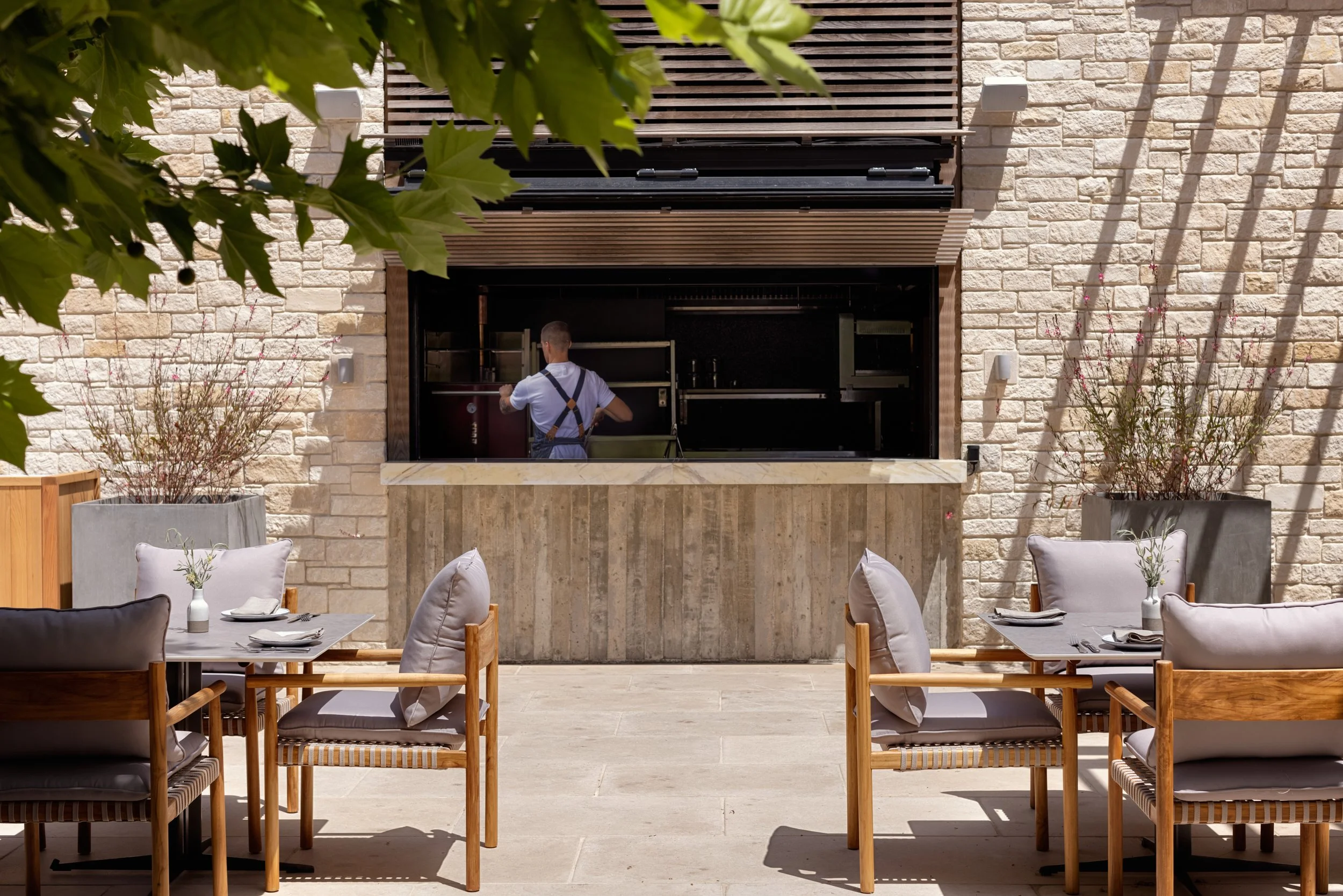 Outdoor dining area with tables set with plates and napkins, facing a stone wall with a built-in outdoor kitchen or bar area, partially shaded by tree branches.