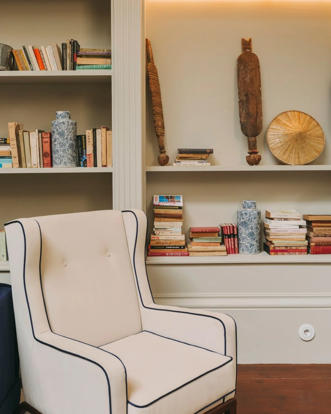 A living room with built-in white bookshelves filled with books, decorative vases, and wooden art pieces. A white armchair with dark piping is in front of the shelves, and a wooden floor is visible.