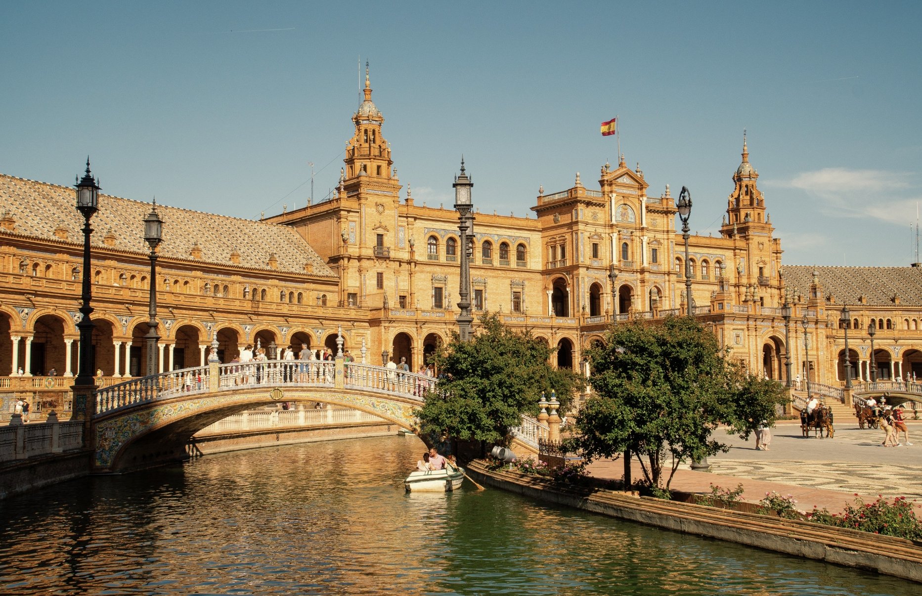 Historic building with arched windows and towers, a flag on top, surrounded by a canal with a small boat and a bridge in the foreground, and people walking and walking horses nearby under a clear sky.