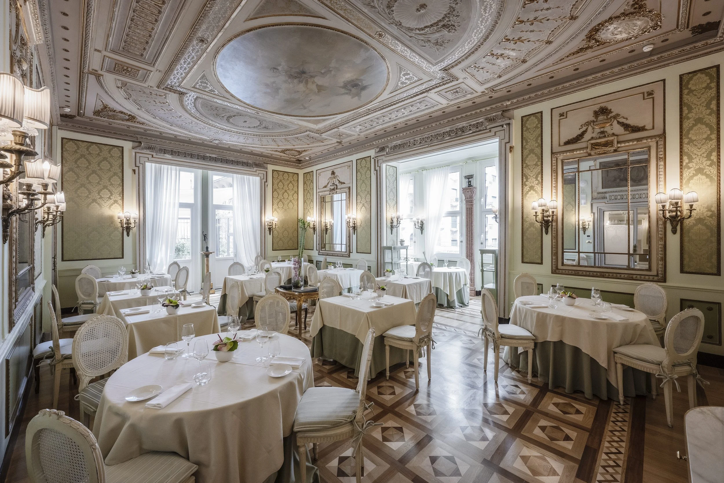 Elegant dining room with high ceilings, intricate ceiling moldings, large windows with white curtains, and decorated walls. Multiple round tables are set with white tablecloths, plates, glasses, and small floral centerpieces. Stylish chairs surround the tables, and the room features antique wall sconces and a polished wooden parquet floor.