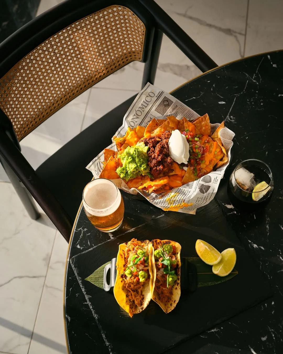 Chips with guacamole, sour cream, ground beef, chopped vegetables, and a drink in a black glass with lime slices, two tacos on a black plate, and a glass of beer on a round black marble table.