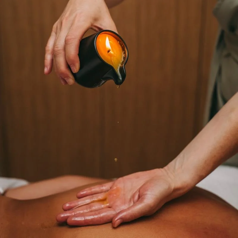 A person pouring warm oil from a black container onto another person's back for massage or spa treatment.