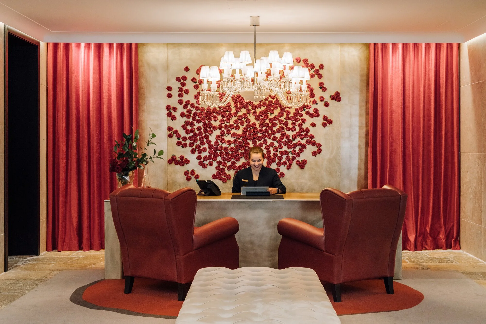 A hotel or reception area with pink curtains, a chandelier, and a decorative wall with red roses. A woman in black uniform is sitting at a desk, and there are pink chairs and a white ottoman in the foreground.