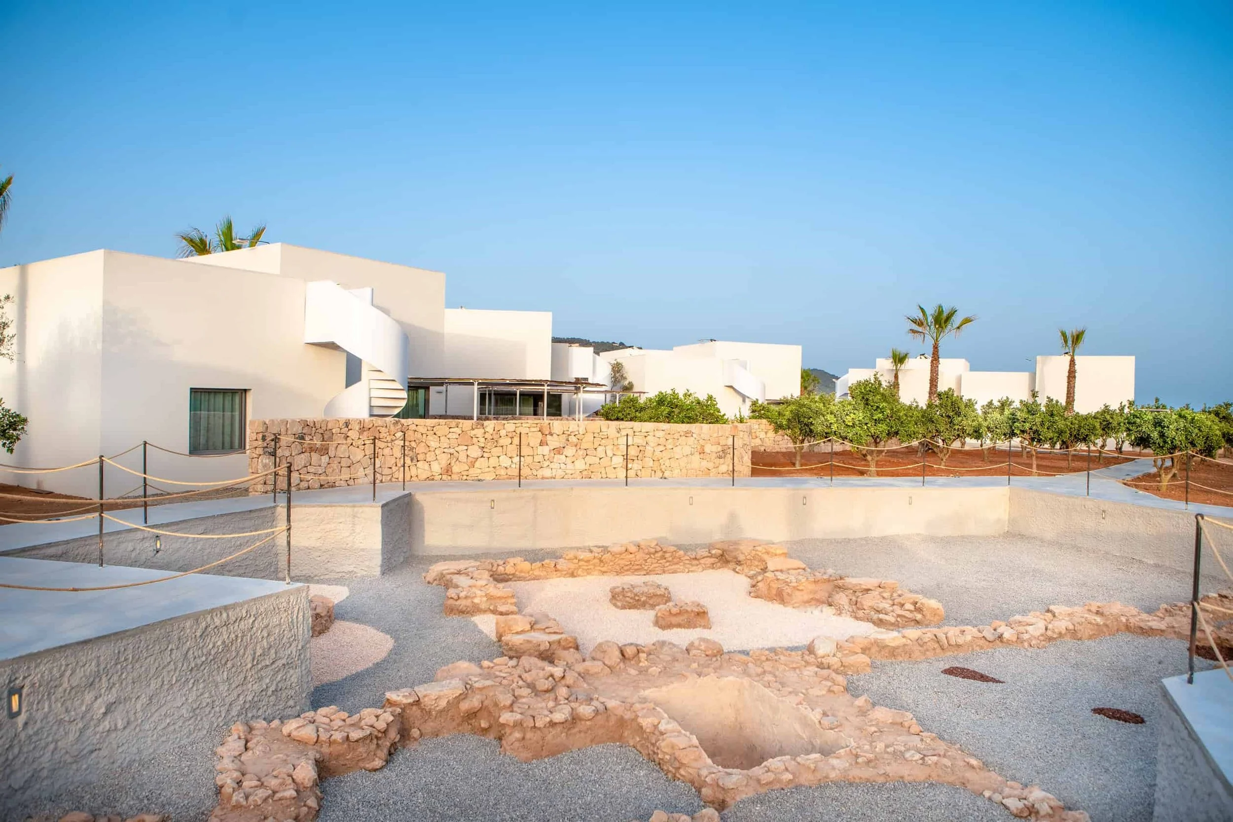 A modern white building with flat roofs, surrounded by a stone wall and a landscaped area with trees and pathways, under a clear blue sky.
