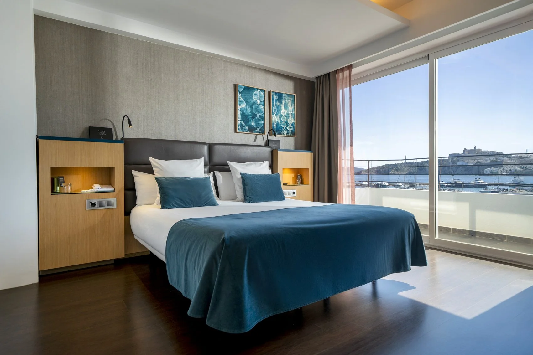 A modern hotel room with a large bed covered in a blue bedspread and matching pillows. The room features a sliding glass door leading to a balcony with a view of boats and a hillside town by the water.