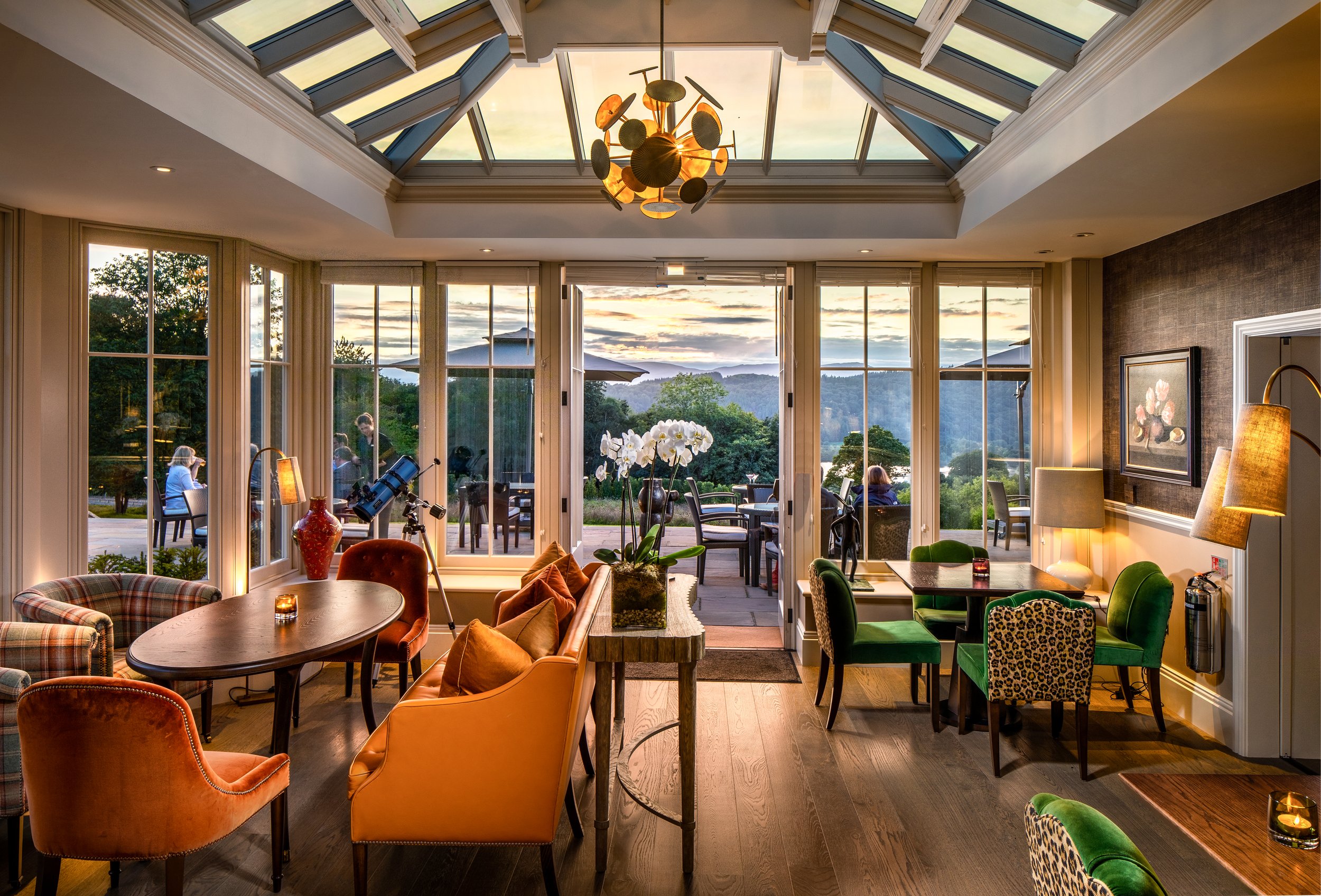 Interior of a cozy living room with large windows and a glass ceiling, showing a scenic outdoor landscape with trees, mountains, and a sunset sky. The room has various chairs, a table, a lamp, and decorative items like a vase of orchids.
