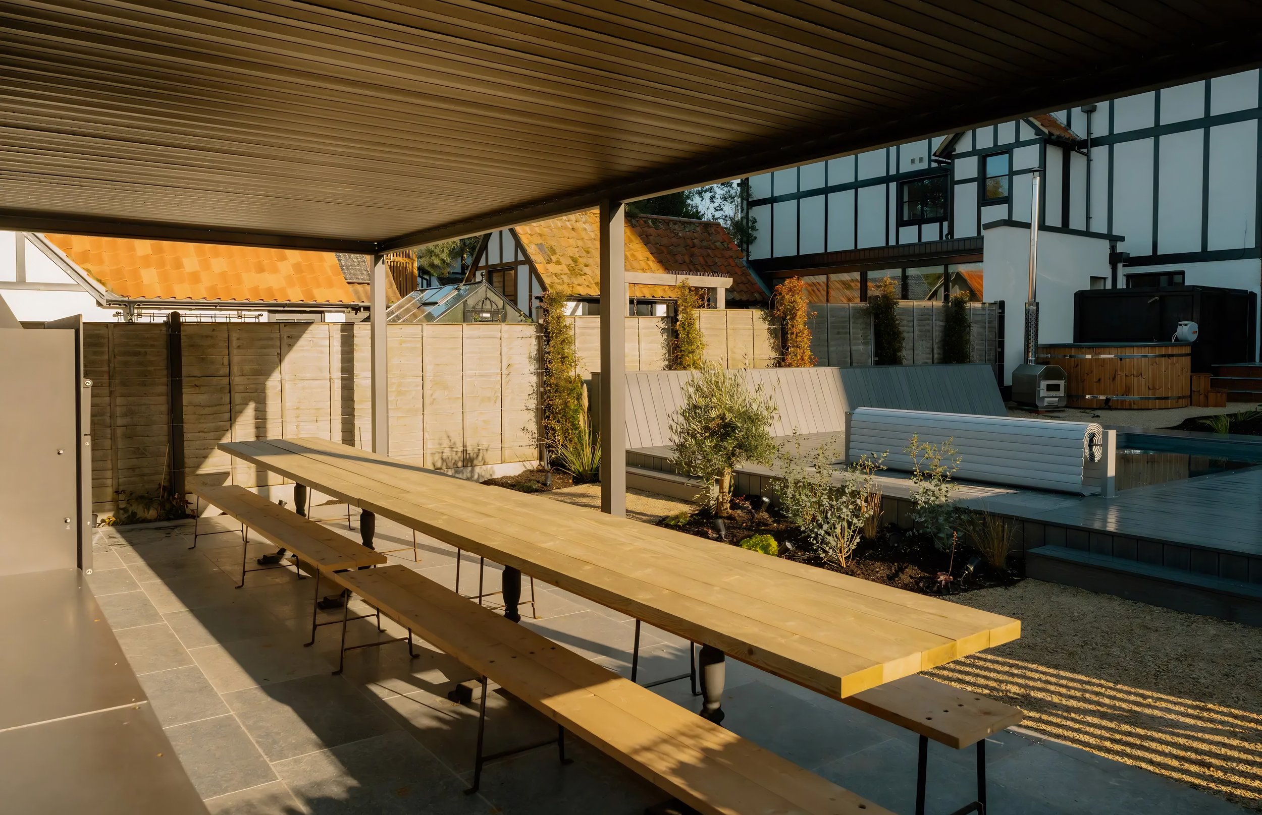 A backyard patio with a wooden picnic table, outdoor seating, and a privacy fence, with houses and trees in the background during daylight.