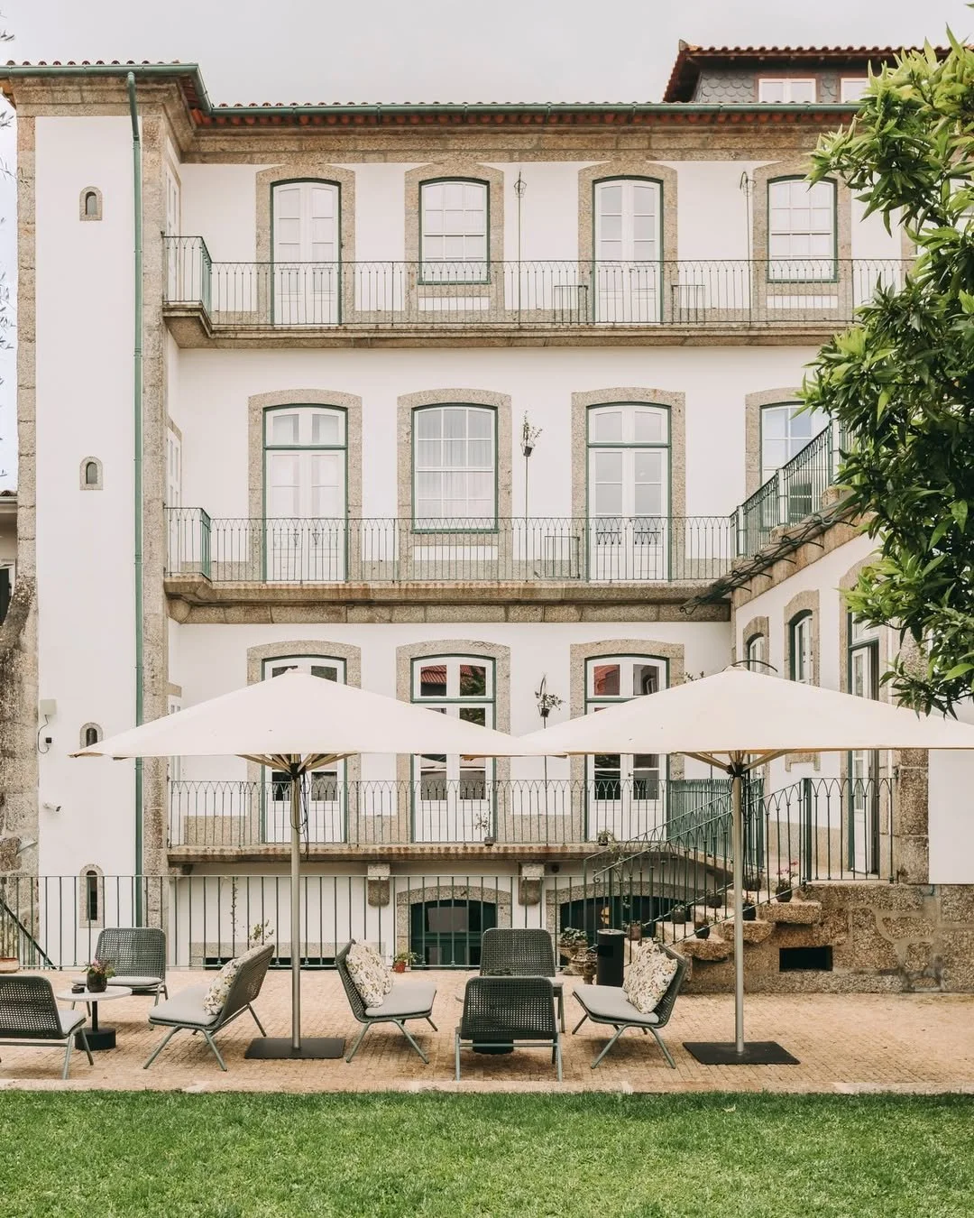 Outdoor patio area with chairs, tables, and umbrellas in front of a multi-story residential building with white walls, stone window frames, and balconies.